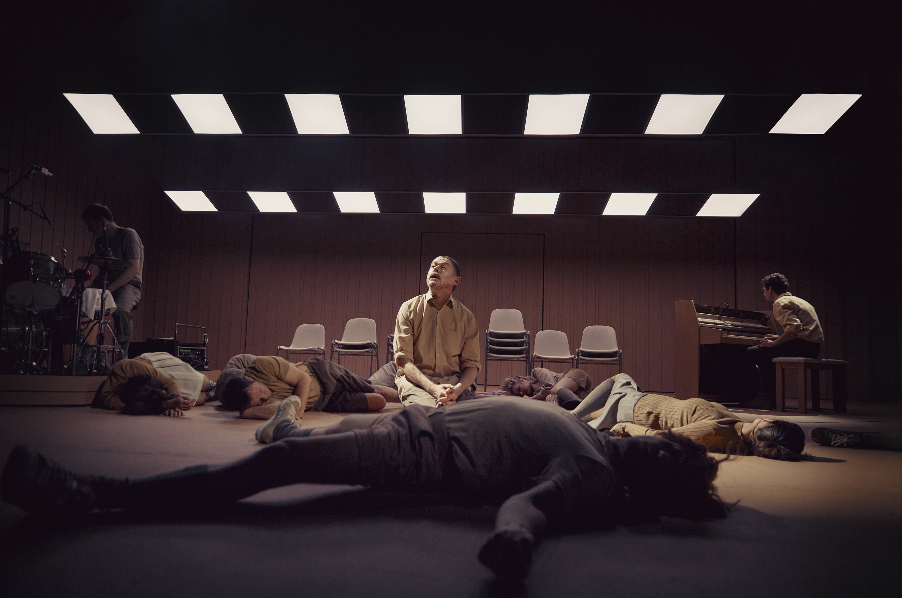 Mand i beige tøj sidder på knæ mellem liggende personer på gulvet. Der sidder en mand ved et trommesæt og en ved et klaver. Lysfelter i loftet. 
