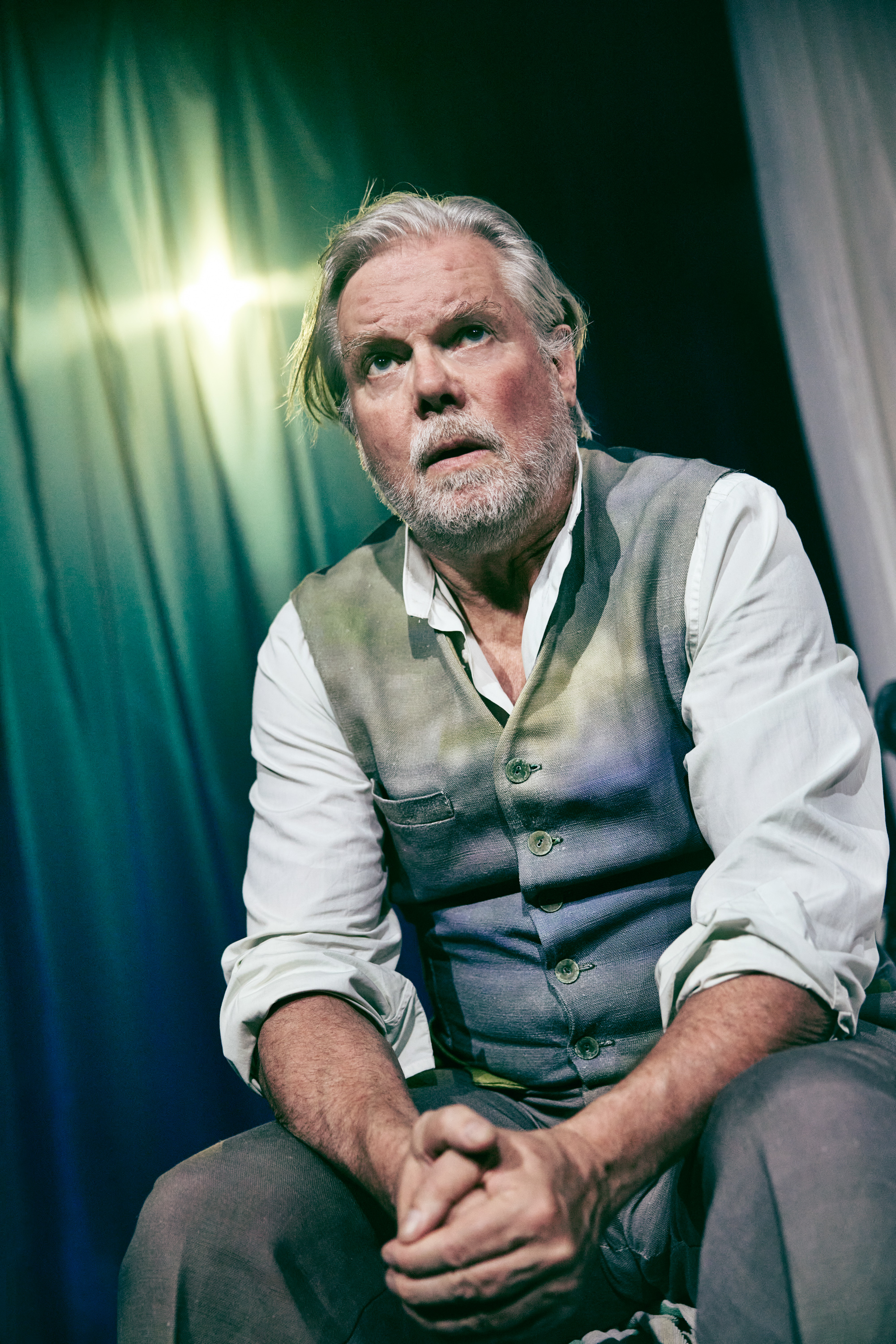 En ældre mand iført hvid skjorte, grå vest og matchende bukser sidder på en stol med foldede hænder. Fra forestillingen Fruen fra havet på Odesense Teater. 