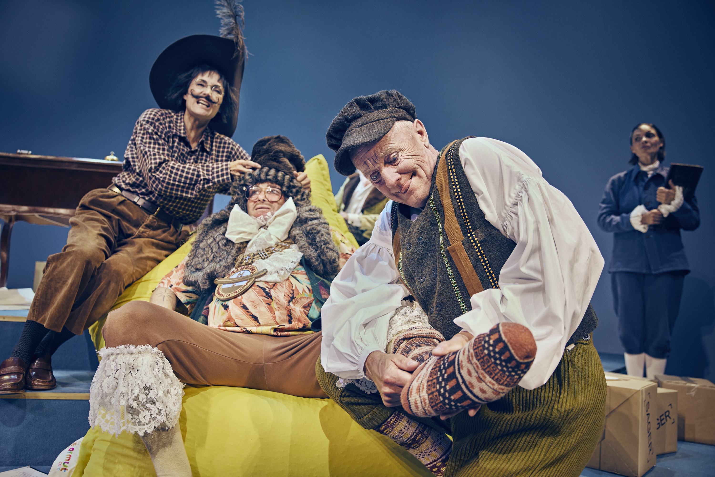 En mand ligger i en sækkestol, mens to andre forsøger at klæde ham på. Fra teaterforestillingen Den politiske kandestøber på Odense Teater