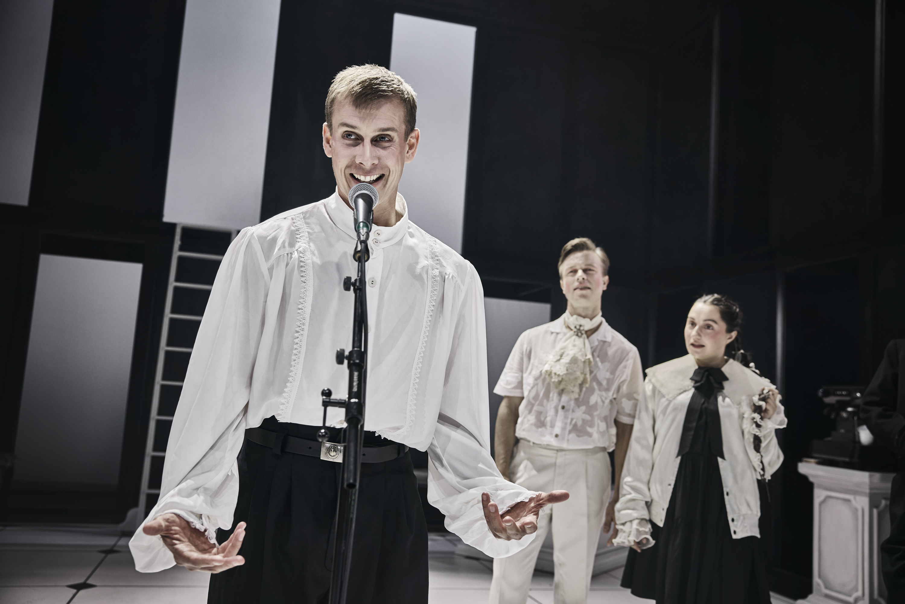 Ung mand i hvid skjorte og sorte bukser står på scenen foran mikrofon. To tilhører stå i baggrunden. Fra forestilling Cyrano de Bergerac på Odense Teater