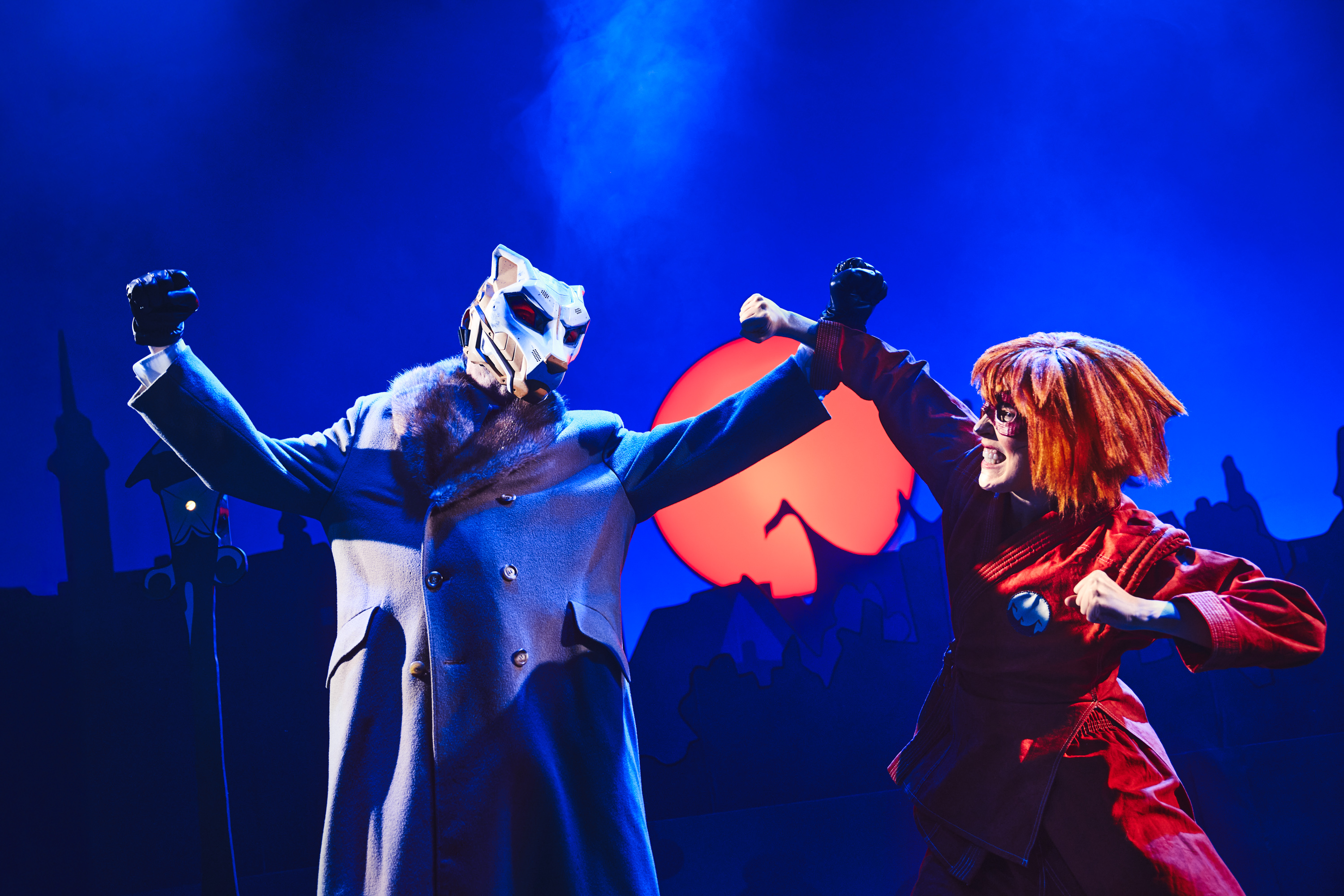 En kampscene mellem en person med ulvemaske og lang frakke mod en pige i rødt kamptøj og heltemaske. Fra forestillingen Håndbog for superhelte på Odense Teater