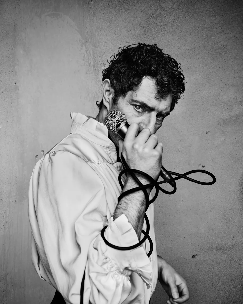 Sort-hvidt foto. Mand med mørkt hår i hvid skjorte med flæser. Holder mikrofon i hånden op mod ansigt, med ledning viklet om hånden.  Cyrano de Bergerac på Odense Teater