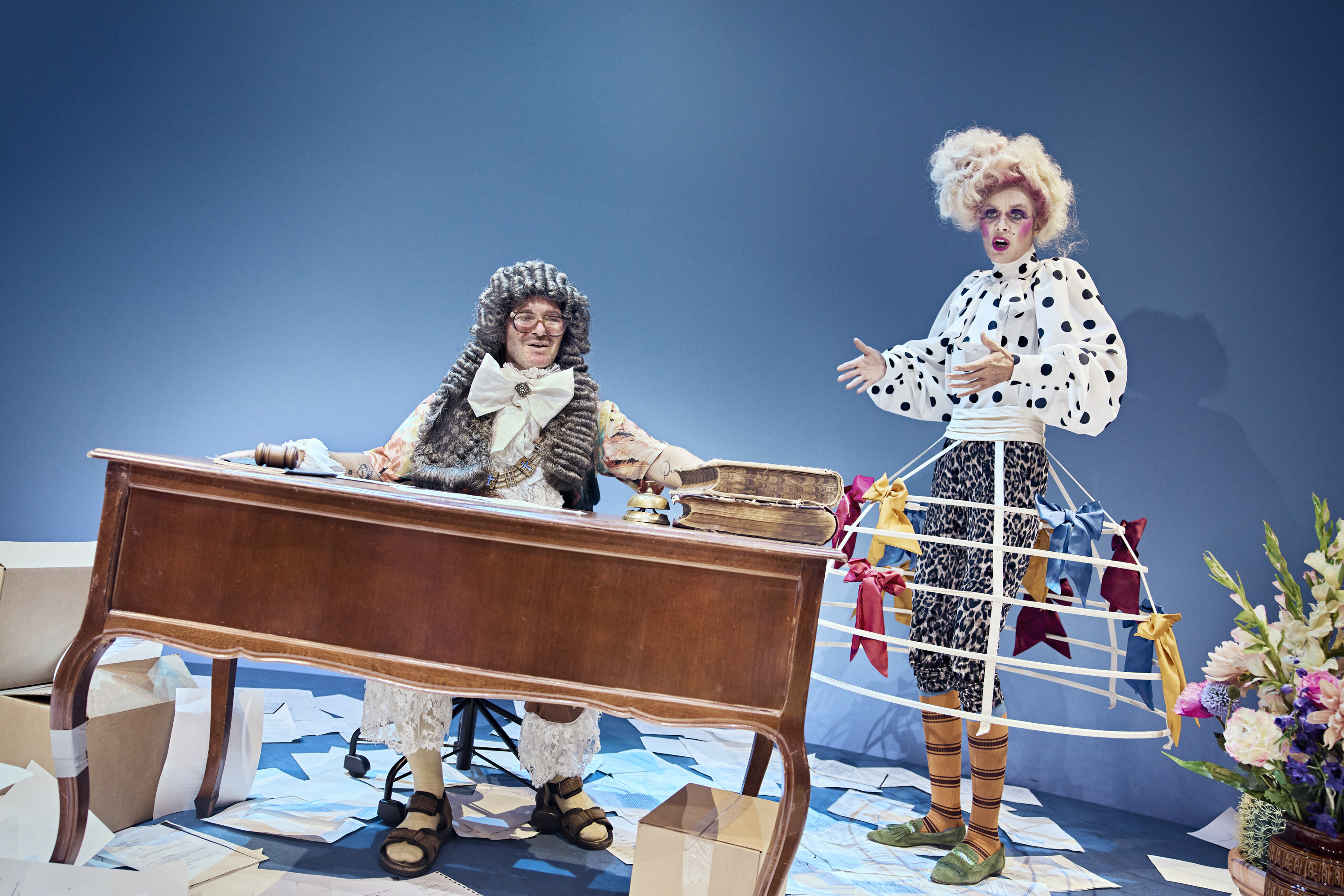 En mands sidder ved et skrivebord. En kvinde med stor paryk og krinoline står ved siden af. Fra teaterforestillingen Den politiske kandestøber på Odense Teater