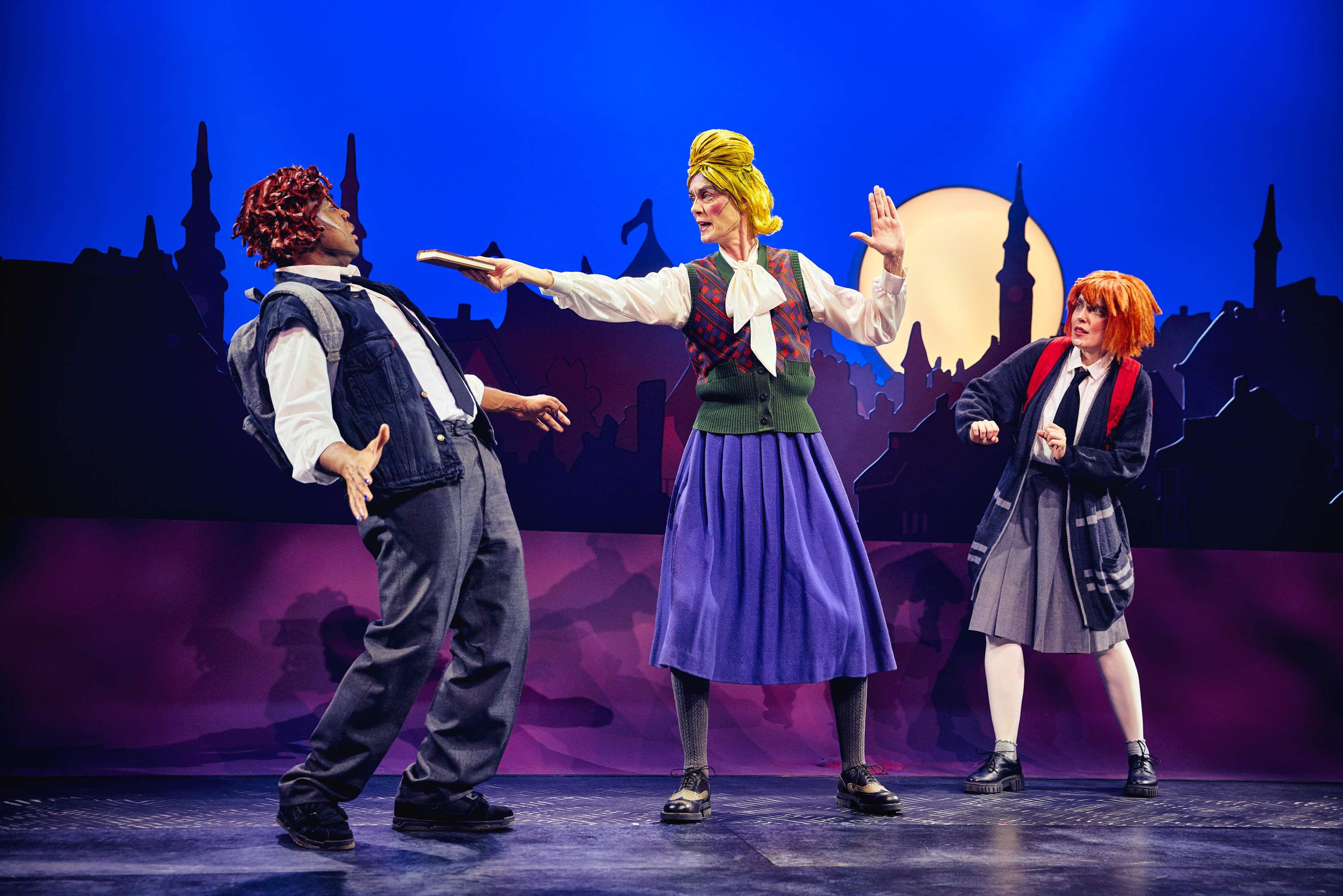 3 personer på en scene. To i skoleuniform og en ældre kvinde med gult hår holder en bog op mod drengen som trækker sig. Fra forestillingen Håndbog for superhelte på Odense Teater