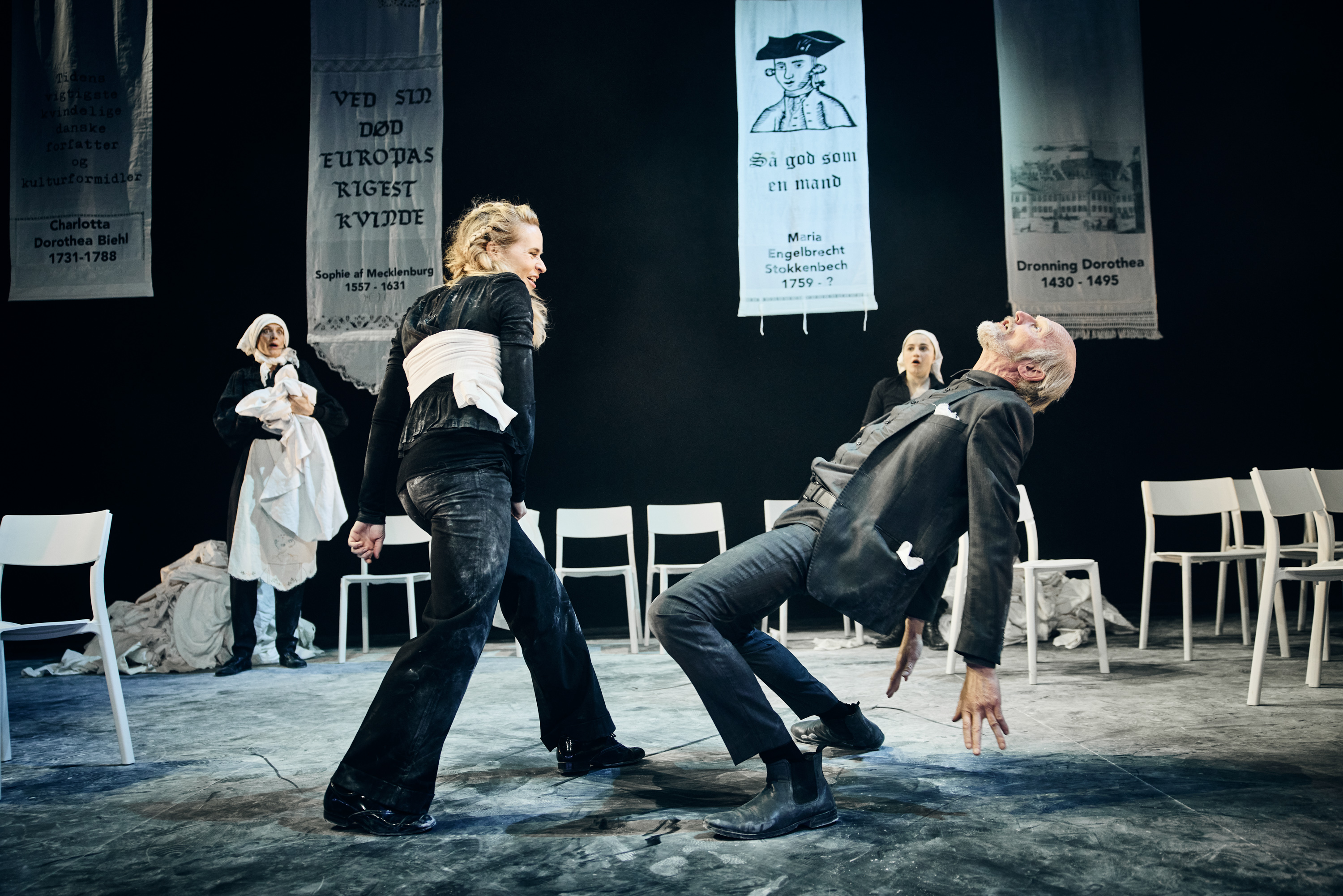 En kvinde klædt i sort. En mand der falder tilbage som er han blevet slået. To kvinder i baggrund ser forskrækket til. Fra forestilling Kvinde kend din historie på Odense Teater