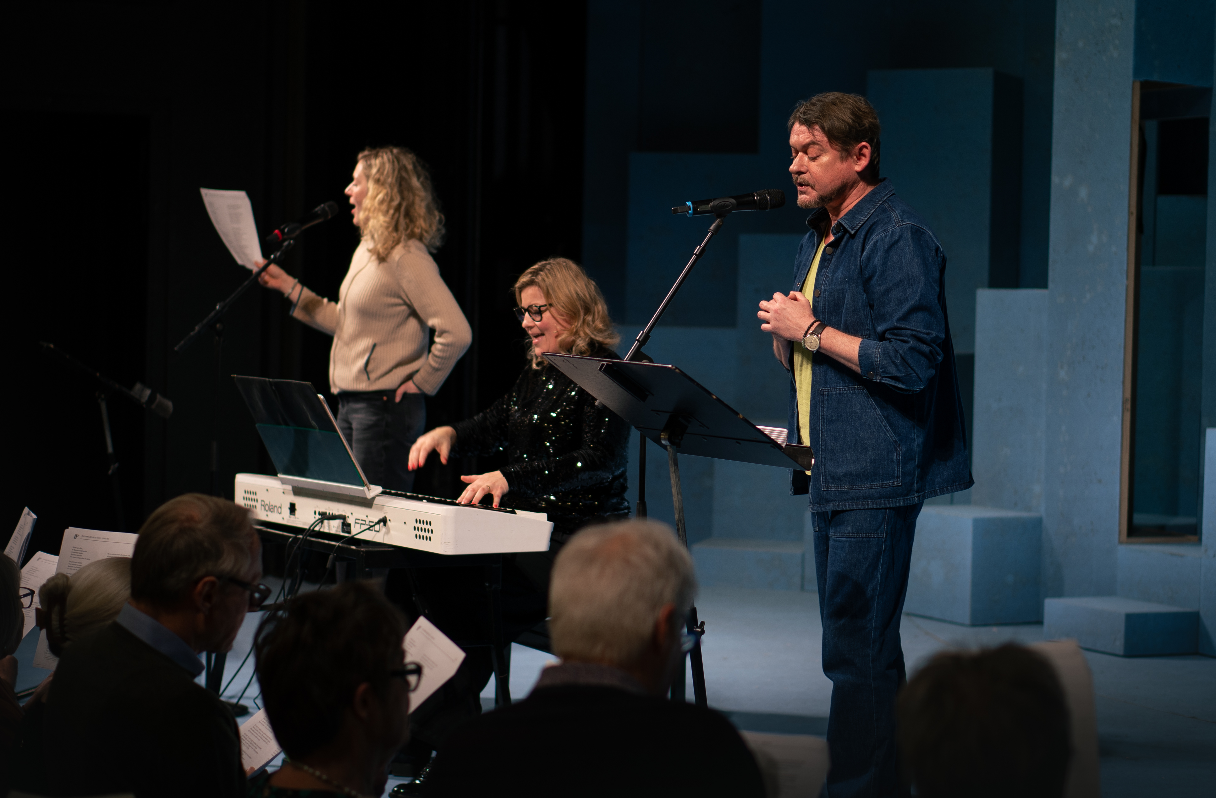 2 kvinder og 1 mand på en scene. Kvinde i midten sidder ved hvidt piano. Mand og kvinde på hver side står ved mikrofon og synger. Publikum anes i front af billedet. 