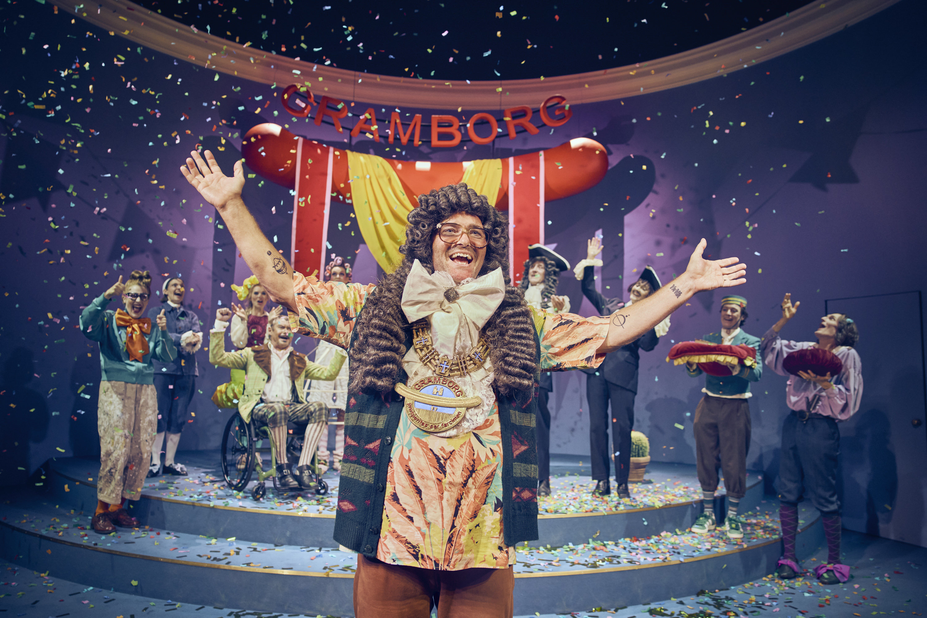 En meget glad og jublende mand slår ud med armene. Konfetti regner ned over ham. I baggrunden jubler flere personer. Fra teaterforestillingen Den politiske kandestøber på Odense Teater