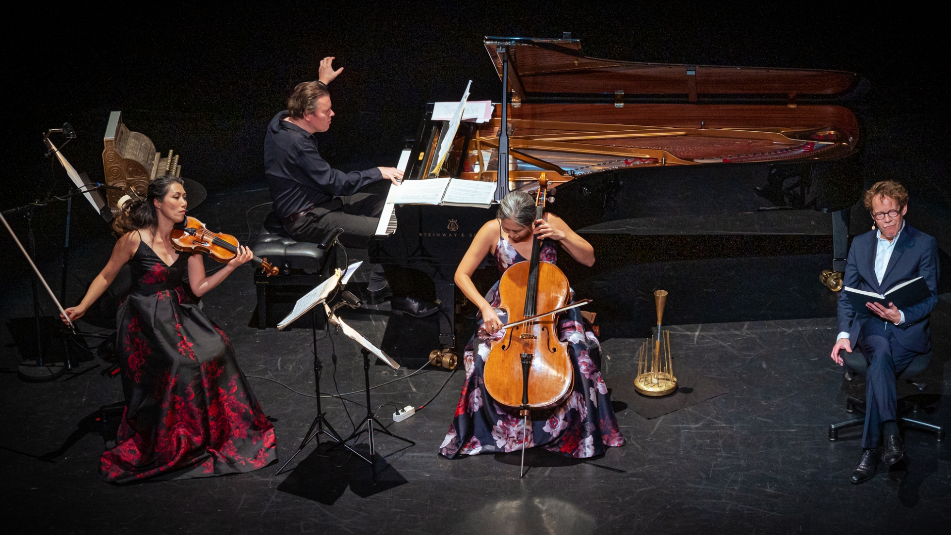Et sort scene og baggrund. tre musikere. En på flygel, en på violin og en på cello. Í højre side sidder en mand med en opslået bog. Fra En digters bazar på Odense Teater