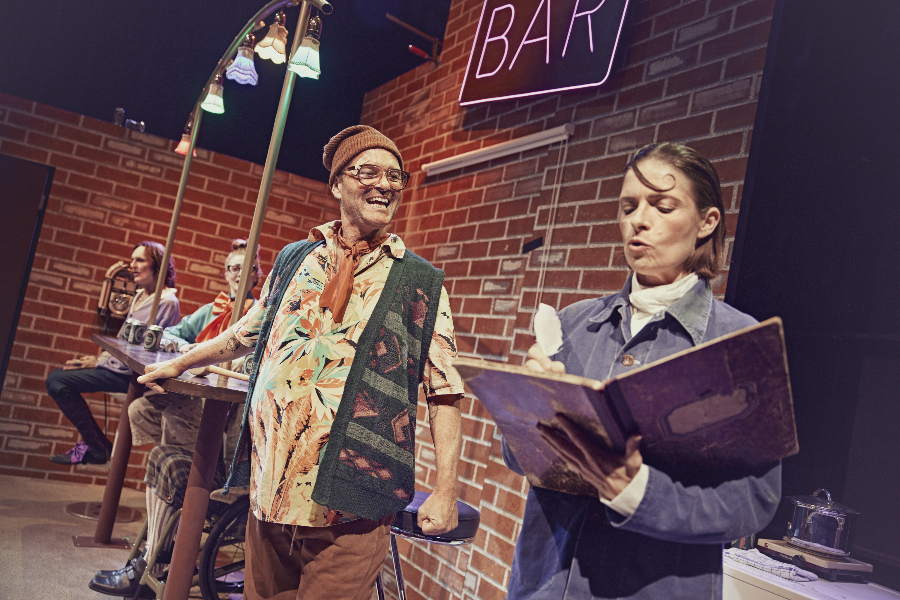 En kvinde skriver noget i en gammel bog. En Mand med strikhue står glad ved siden af. Fra teaterforestillingen Den politiske kandestøber på Odense Teater