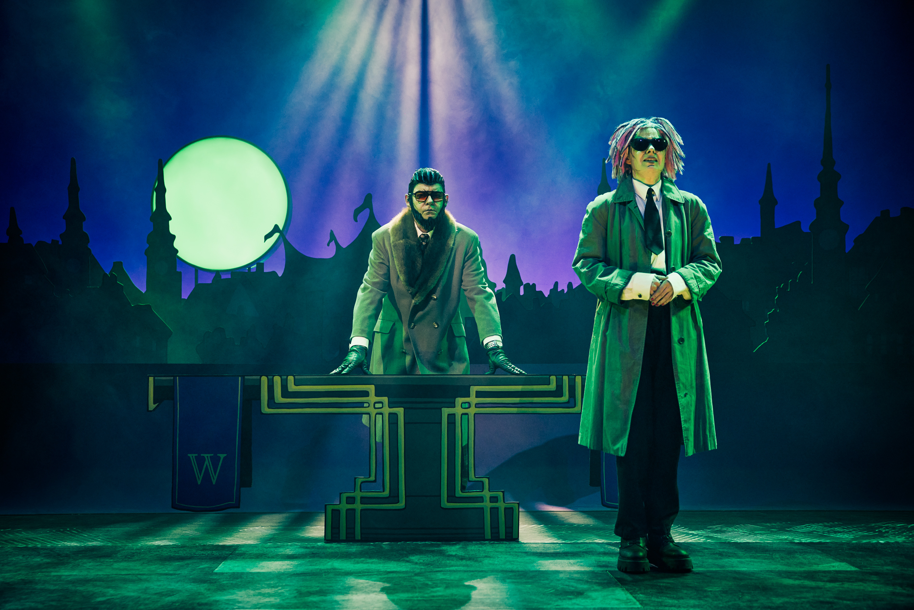 To personer på en scene. De bærer solbriller. Stemning er mørk. En stor fuldmåne i baggrunden. Fra forestillingen Håndbog for superhelte på Odense Teater