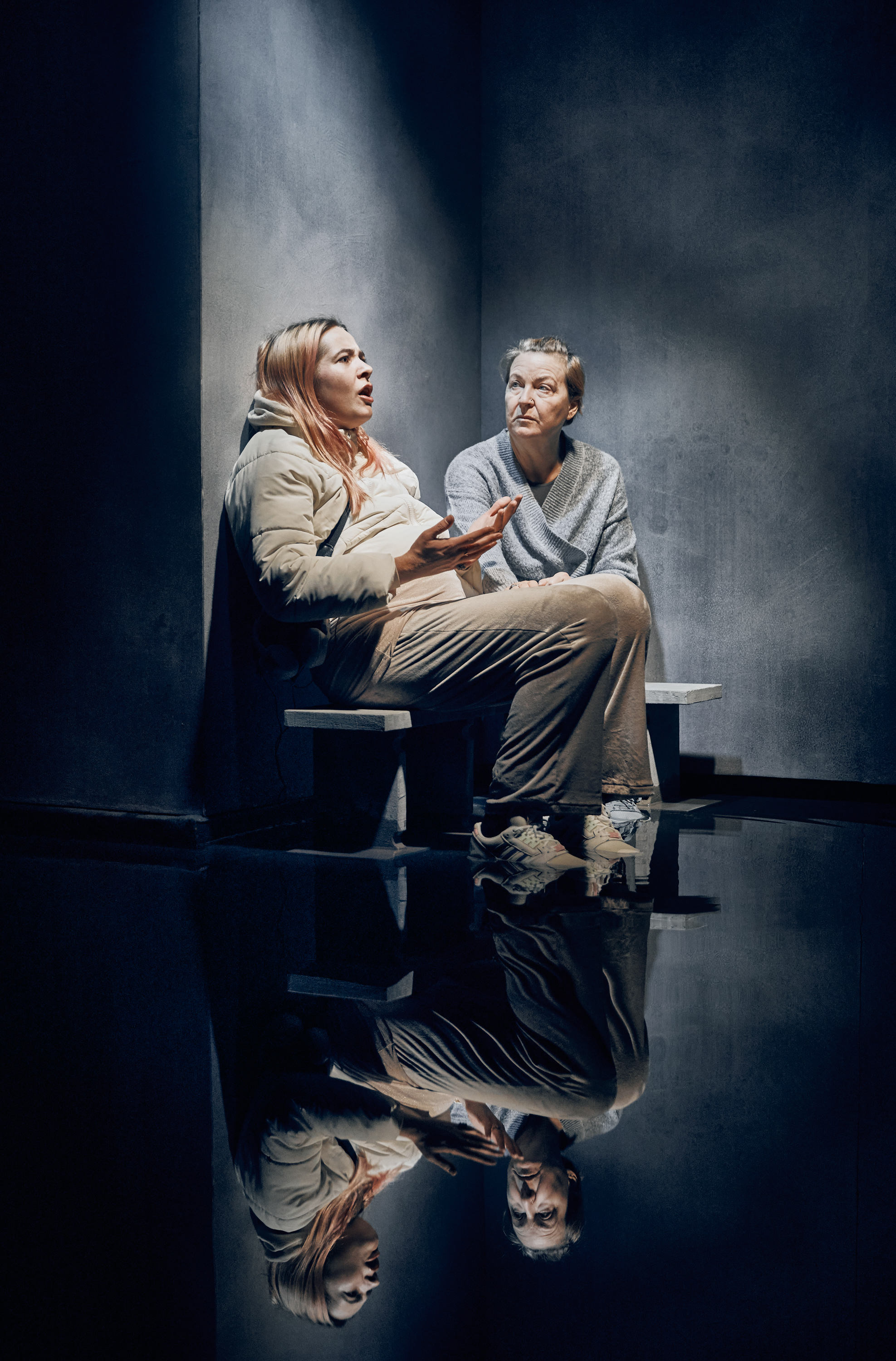 2 kvinder sidder på bænk. Den ene taler. Den anden lytter. Deres kroppe spejler sig i gulvet. Fra forestilling Maria på Odense Teater.