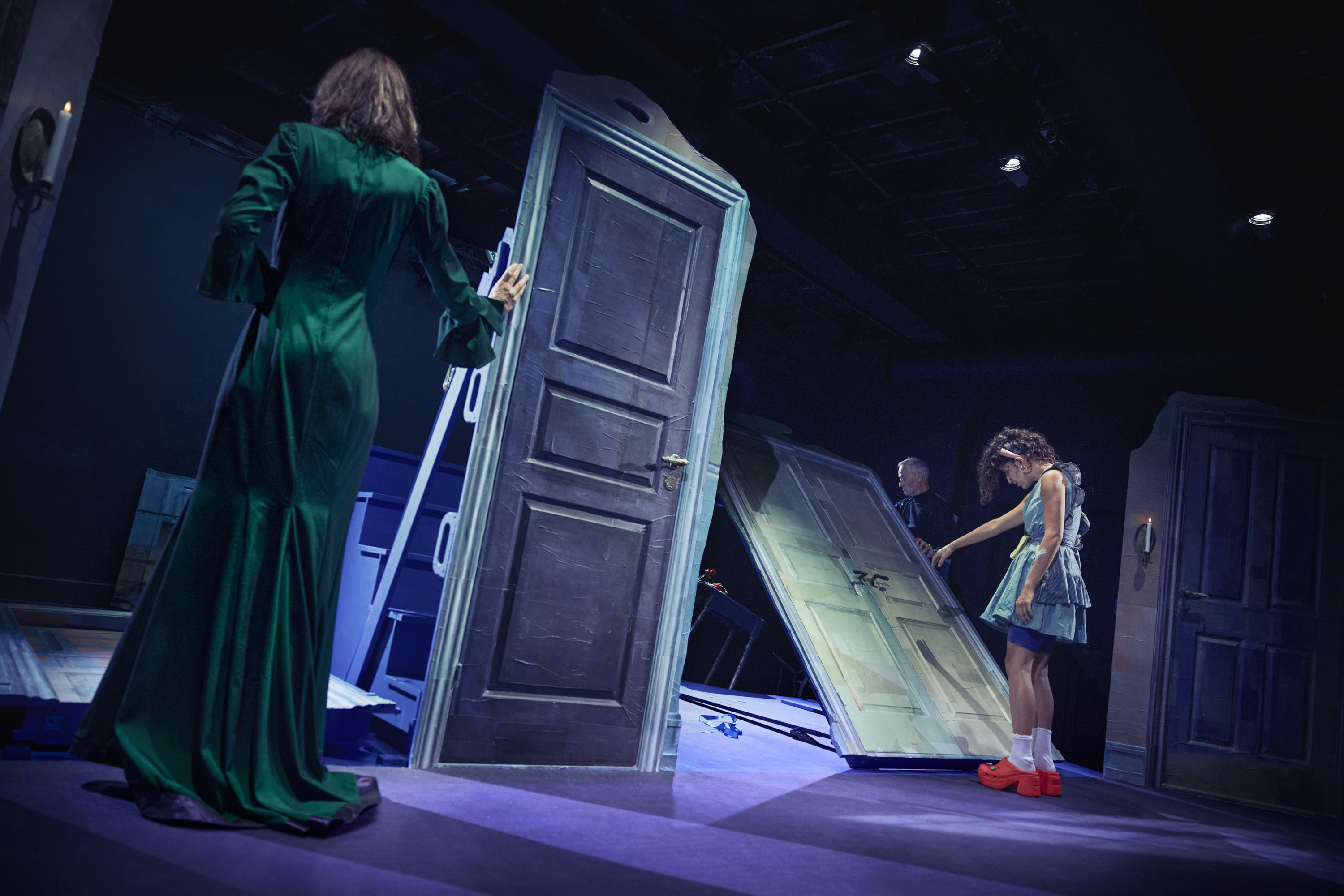 tre skuespillere på scene i scenografi med døre der vælter. Høj kvinde i lang grøn kjole. En kvinde i kort kjole med kraftige orange sko.