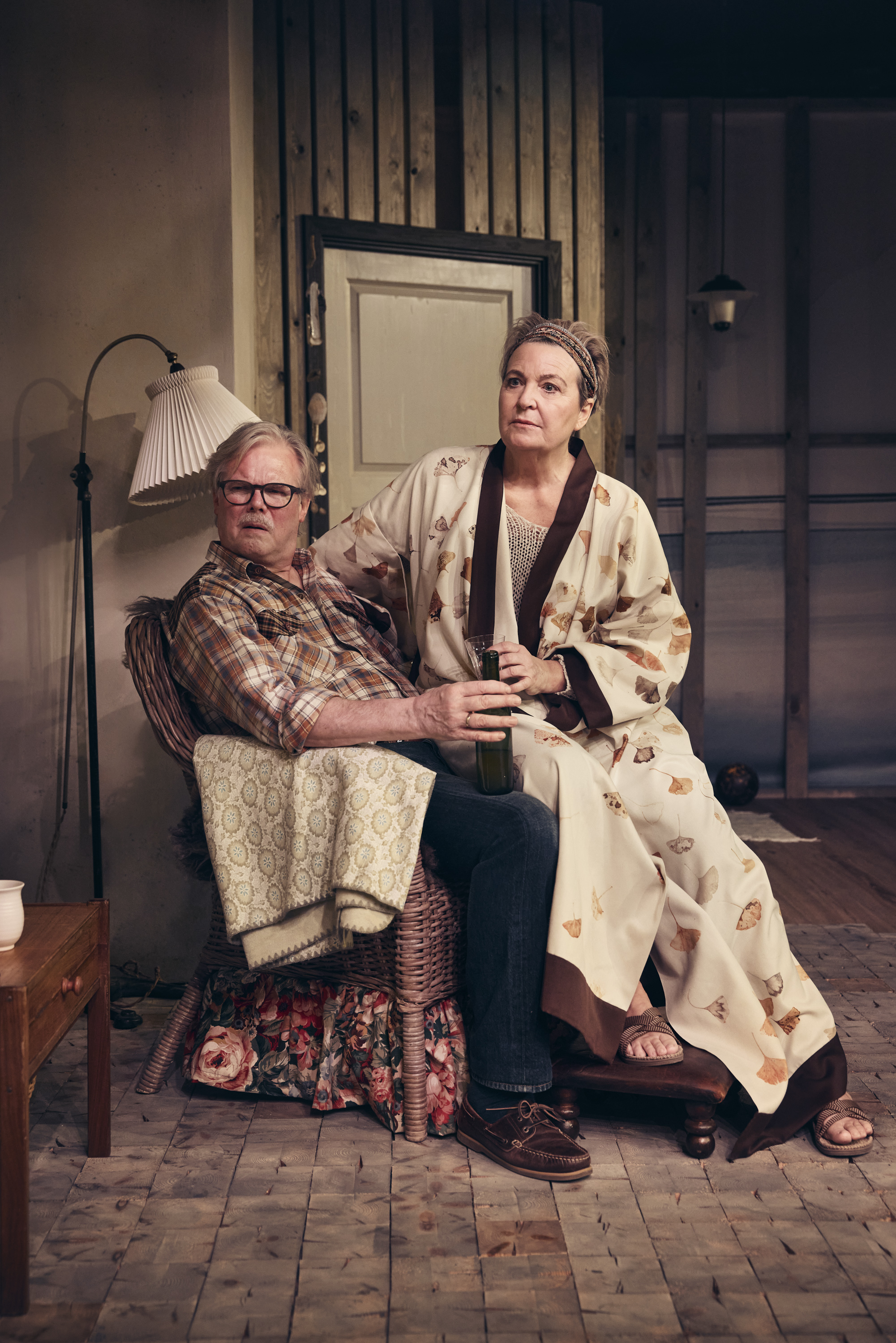 Ældre mand med mørke briller sidder i lænestol med kvinde i kimono på skødet. Fra teaterforestilling Børnene på Odense Teater.