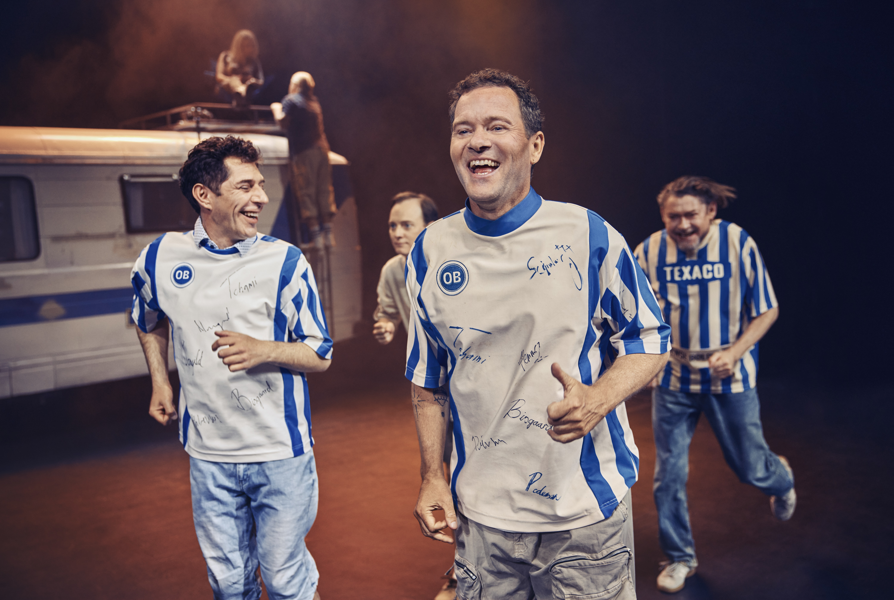 Fire mænd i OB fodboldtrøjer løber i mens de griner. Fra teaterforestillingen Miraklet i Madrid på Odense Teater