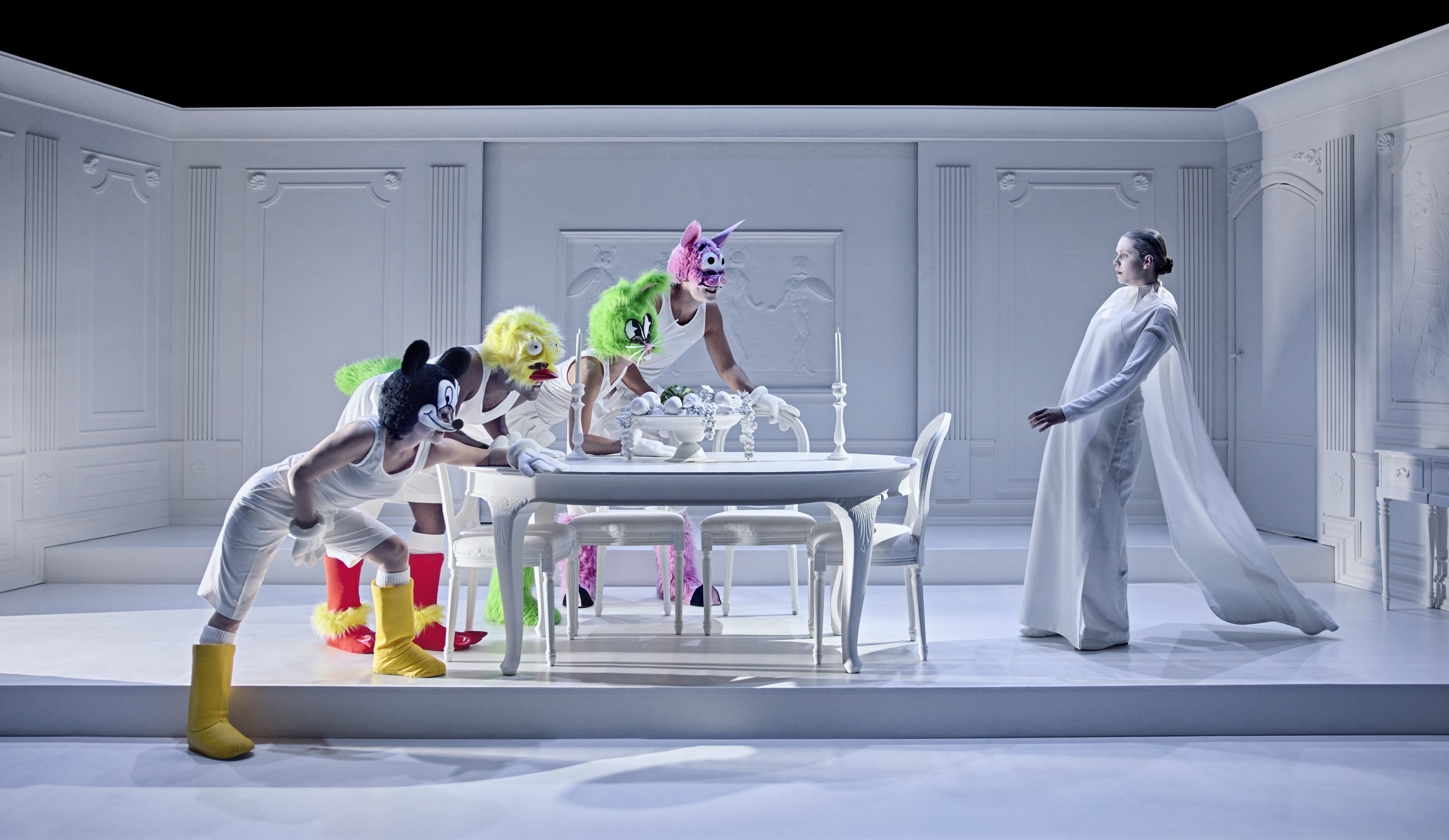 Hvidt senerum. Tegneseriefigurer står lænet ind over hvidt bord. En kvinde i hvidt står for bordenden. Fra forestilling Resten kender du fra film på Odense Teater