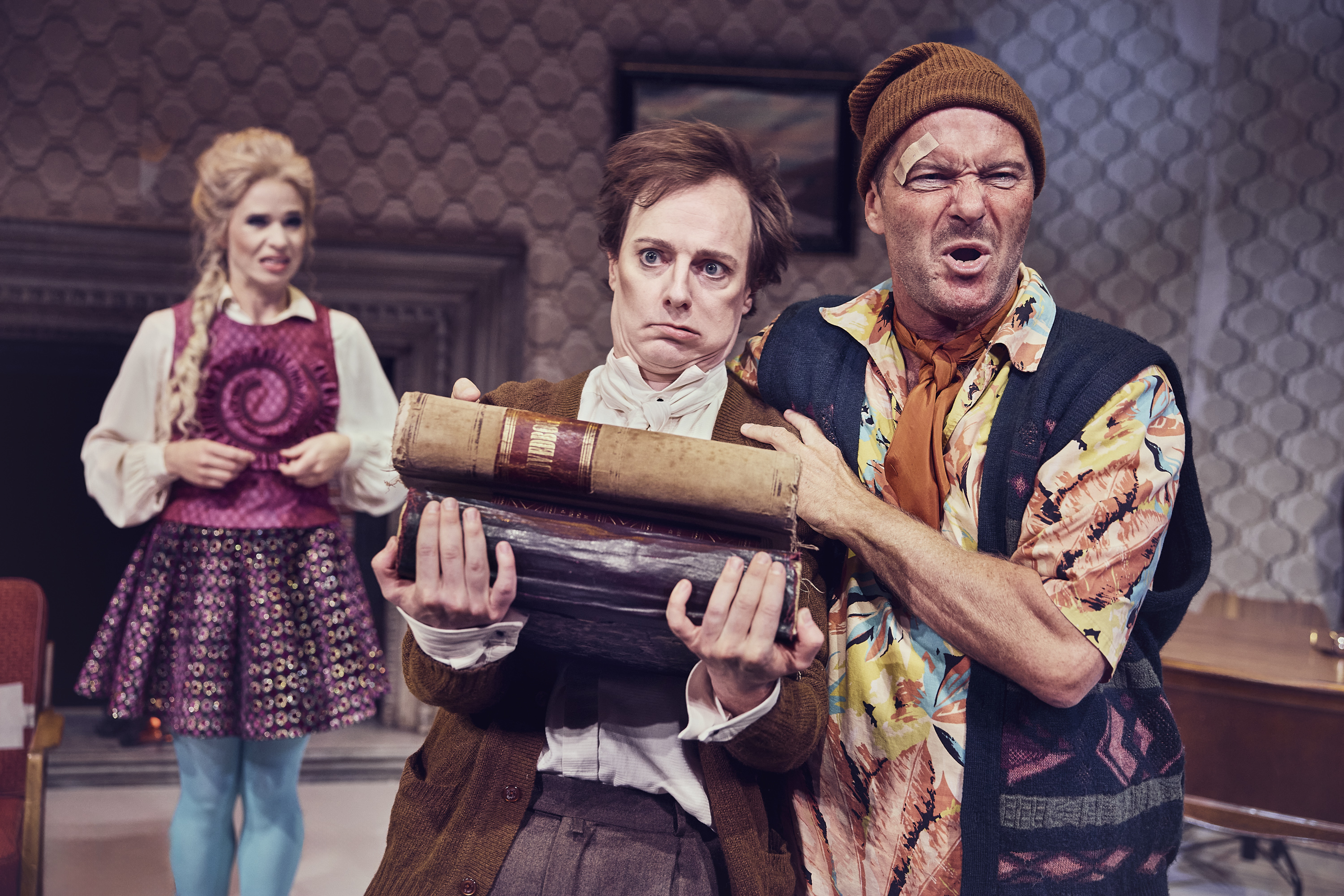 En ung mand får læsset tunge bøger op i armene af en anden mand. En kvinde står i baggrunden. Fra teaterforestillingen Den politiske kandestøber på Odense Teater