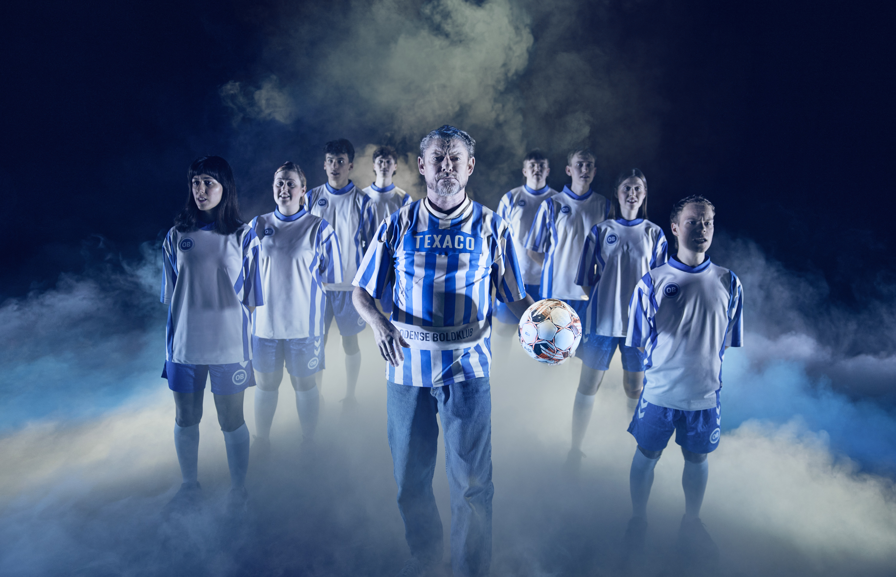 En mand i stribet fodboldtrøje går forrest i formation otte andre fodboldspillere. Fra forestillingen Miraklet i Madrid på Odense Teater.