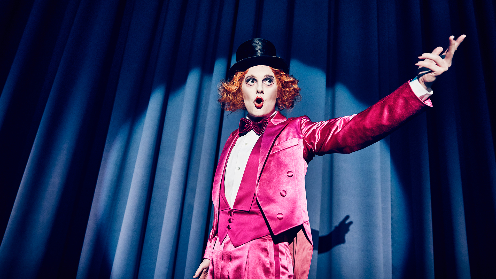 En person på scene med blåt bagtæppe iført stæk pink smoking og butterfly, lg med høj sort hat og orange paryk. 