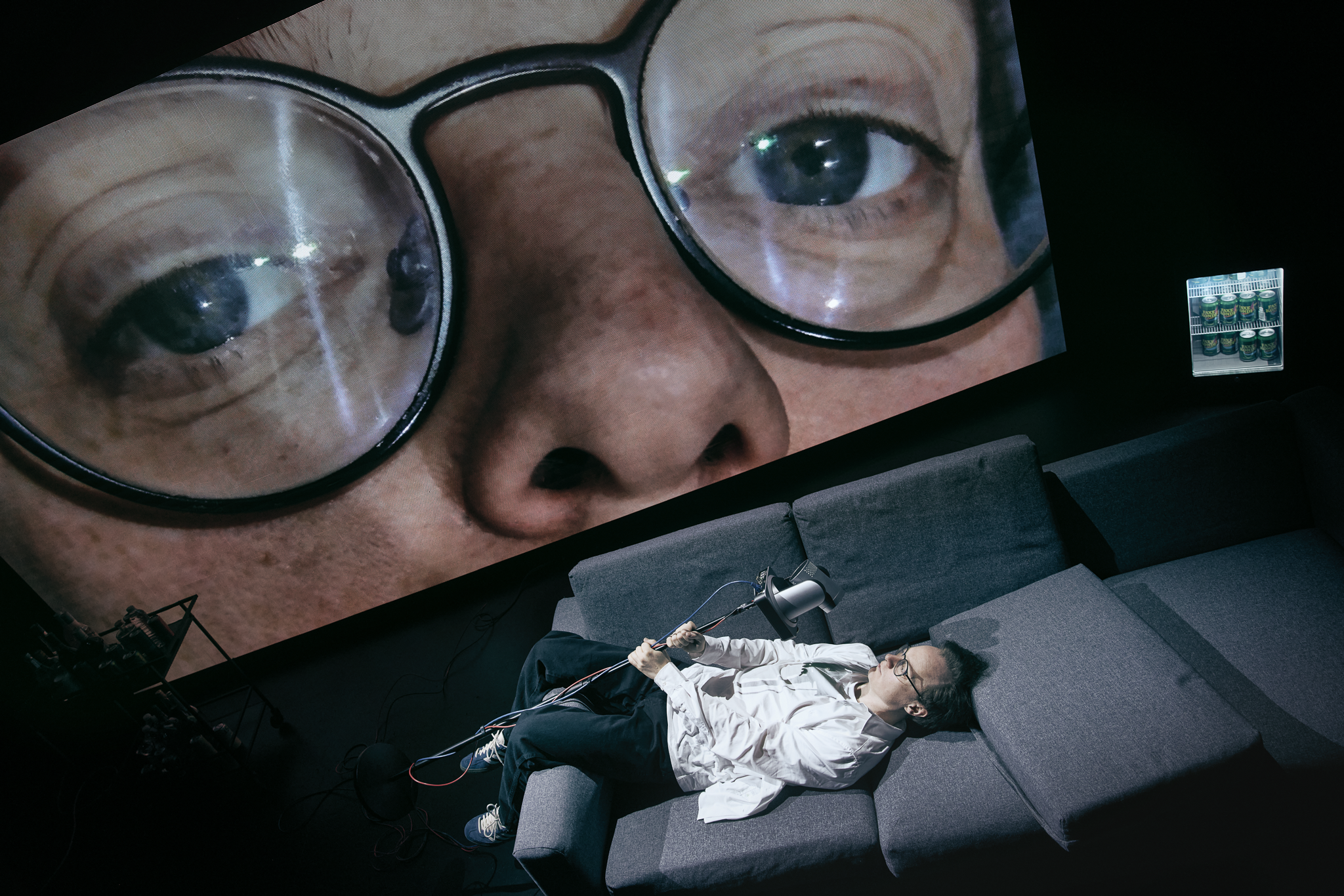 En mand ligger på ryggen på en sofa. Holder en stang med et kamera på som filmer hans ansigt. Bag ham er en stor skærm med nærbillede af hans ansigt. Fra forestillingen AI know you på Odense Teater
