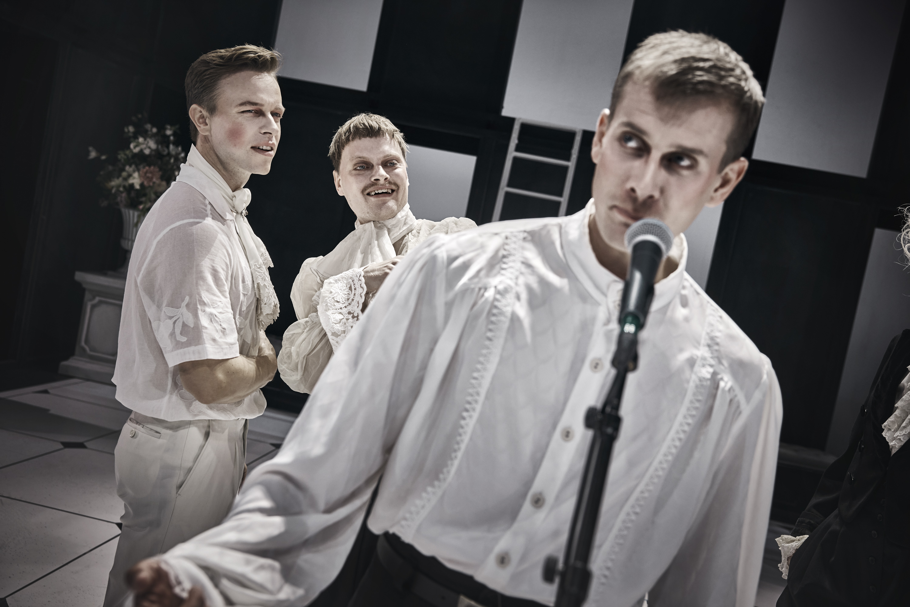 Ung mand i hvid skjorte står på scenen foran mikrofon. To tilhører stå i baggrunden. Fra forestilling Cyrano de Bergerac på Odense Teater