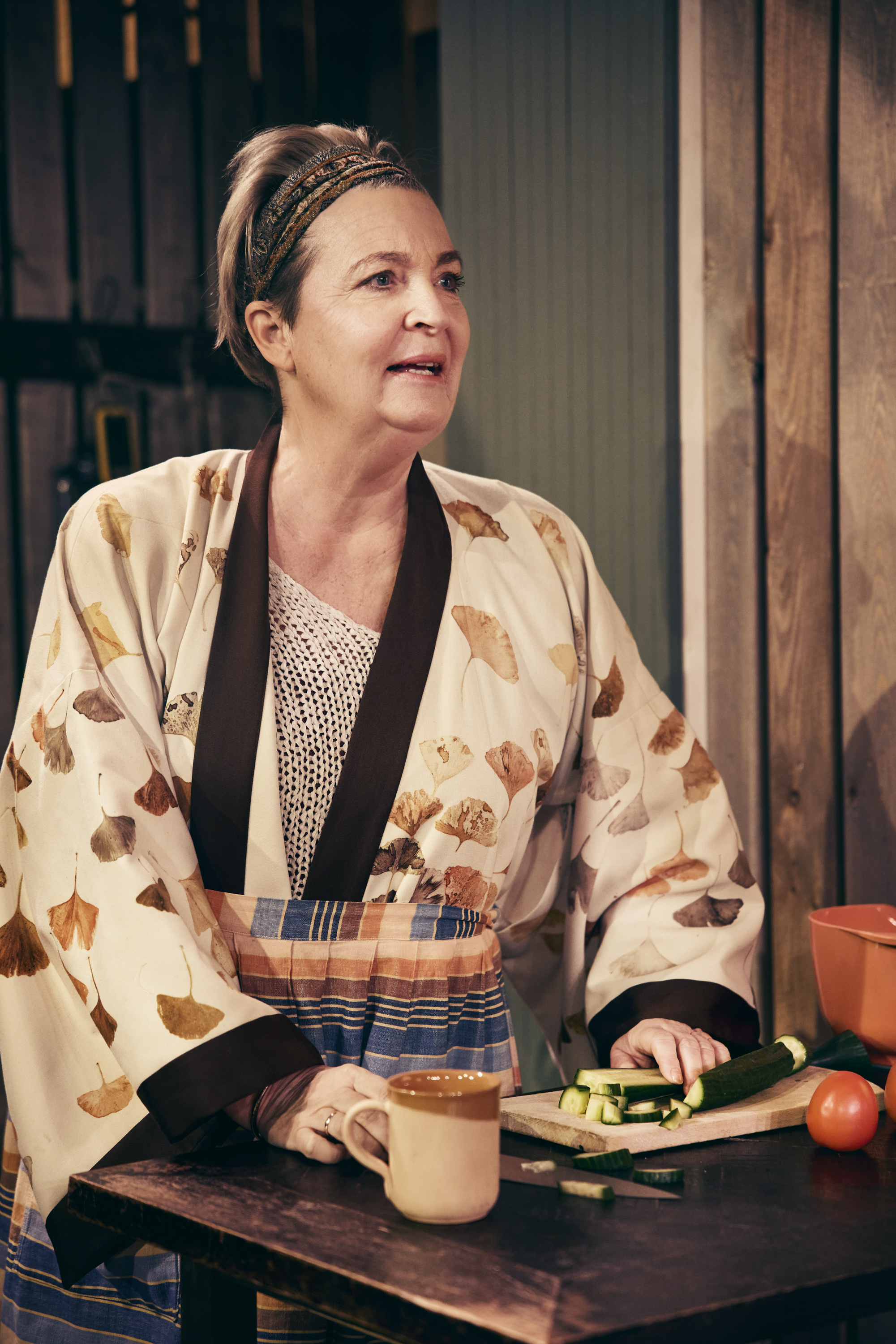 Korthåret kvinde med tørklæde iklædt kimono, står ved køkkenbord og skærer agurk. Fra teaterforestilling Børnene på Odense Teater.