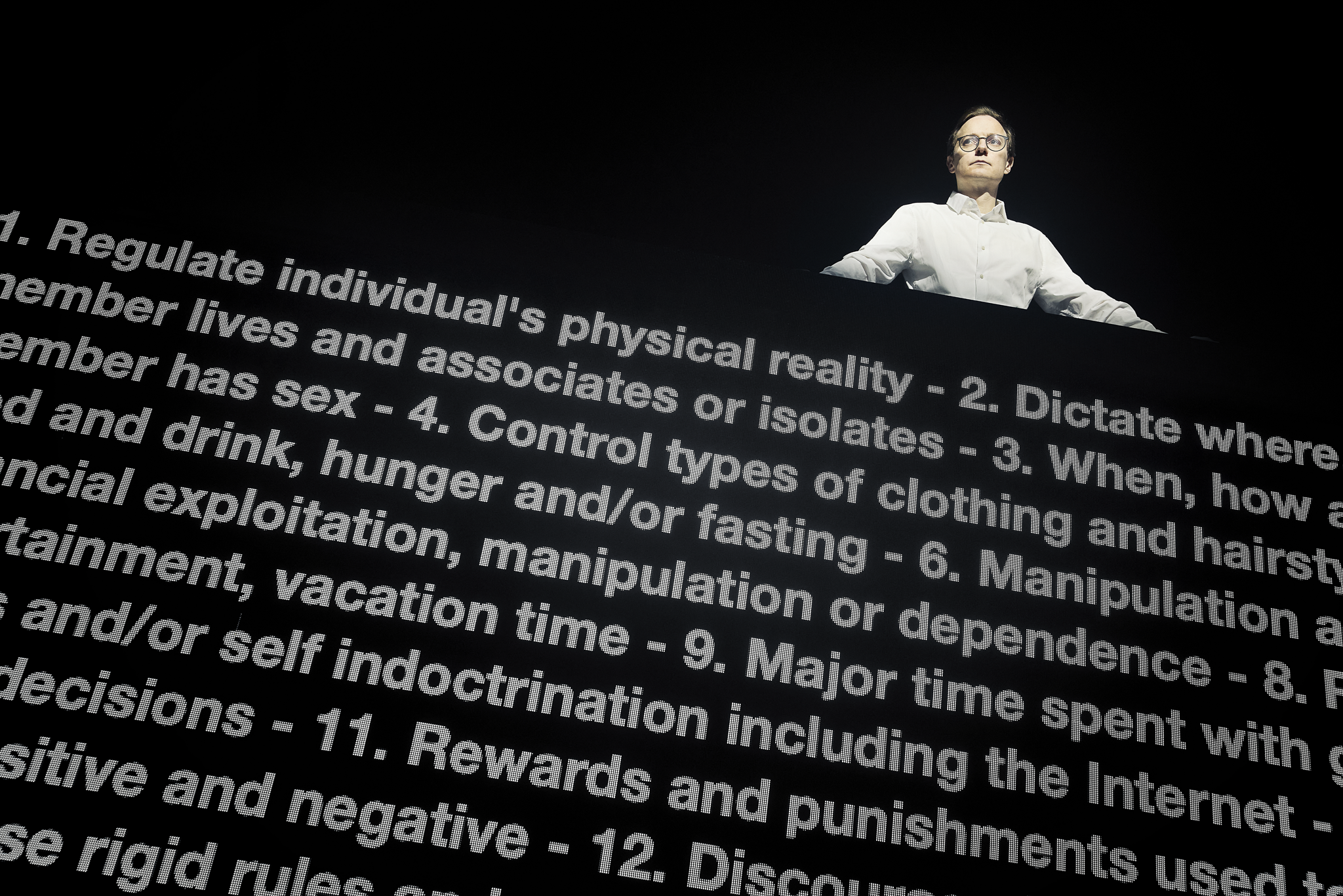 En mand i hvid skjorte står bag en meget stor skærm med en masse hvid tekst.. Man kan kun se hans overkrop. Fra forestillingen AI know you på Odense Teater