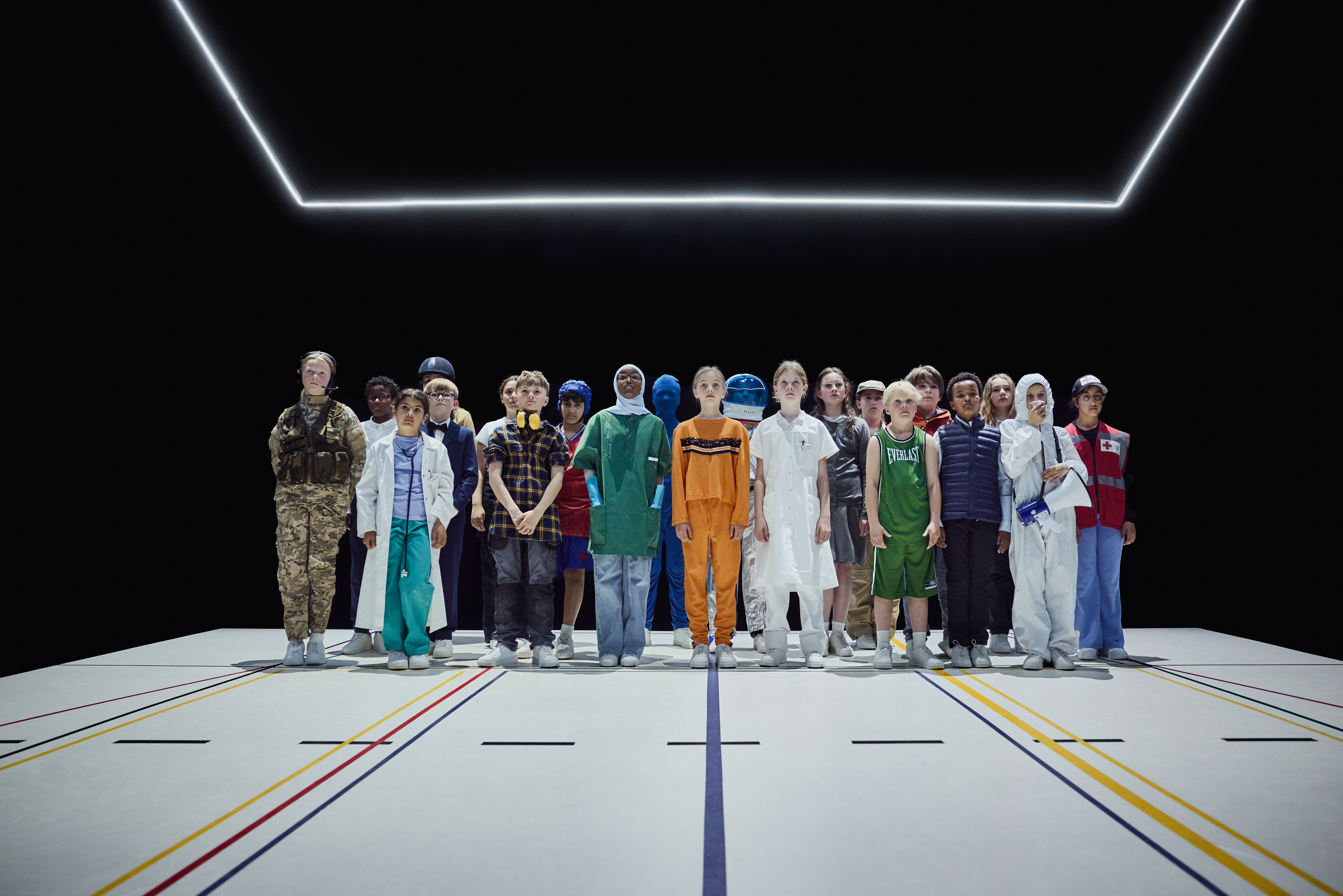 En gruppe af børn i forskellige arbejdsrelaterede outfits står i sort rum på hvidt gulv. Fra forestillingen Mod alle odds på Odense Teater