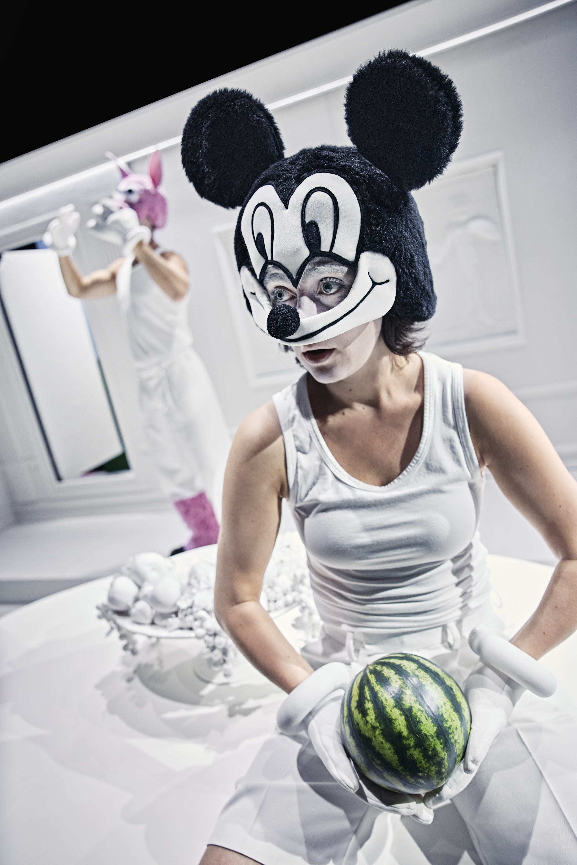 En kvinde i hvid undertrøje og shorts med stor Mickie Mouse maske og hvide handsker holder en grøn vandmelon. Fra forestilling Resten kender du fra film på Odense Teater