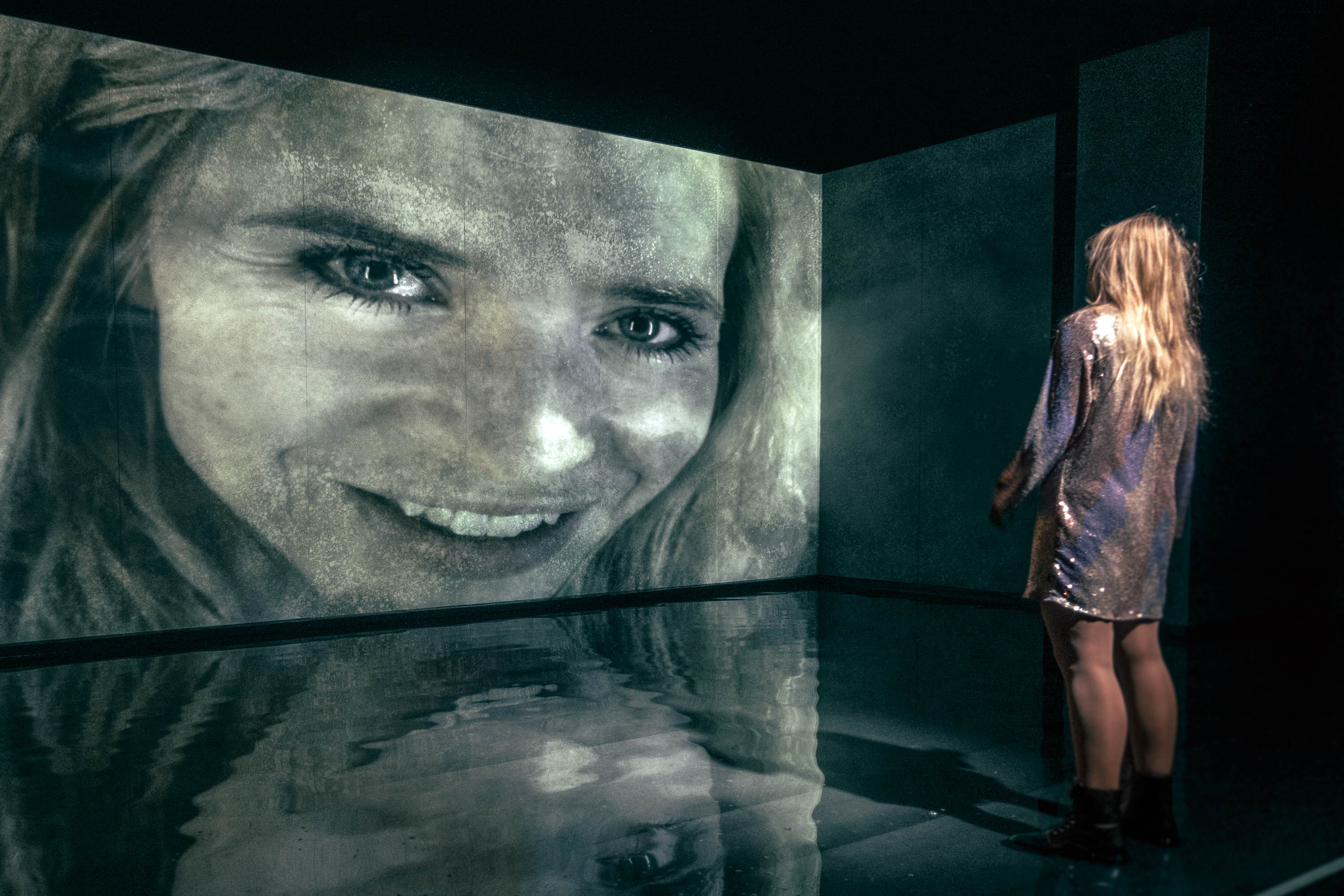 Ung kvinde i pailletkjole står med front mod stor skærm, hvorpå der er et smilende kvindeansigt. Fra forestilling Maria på Odense Teater.