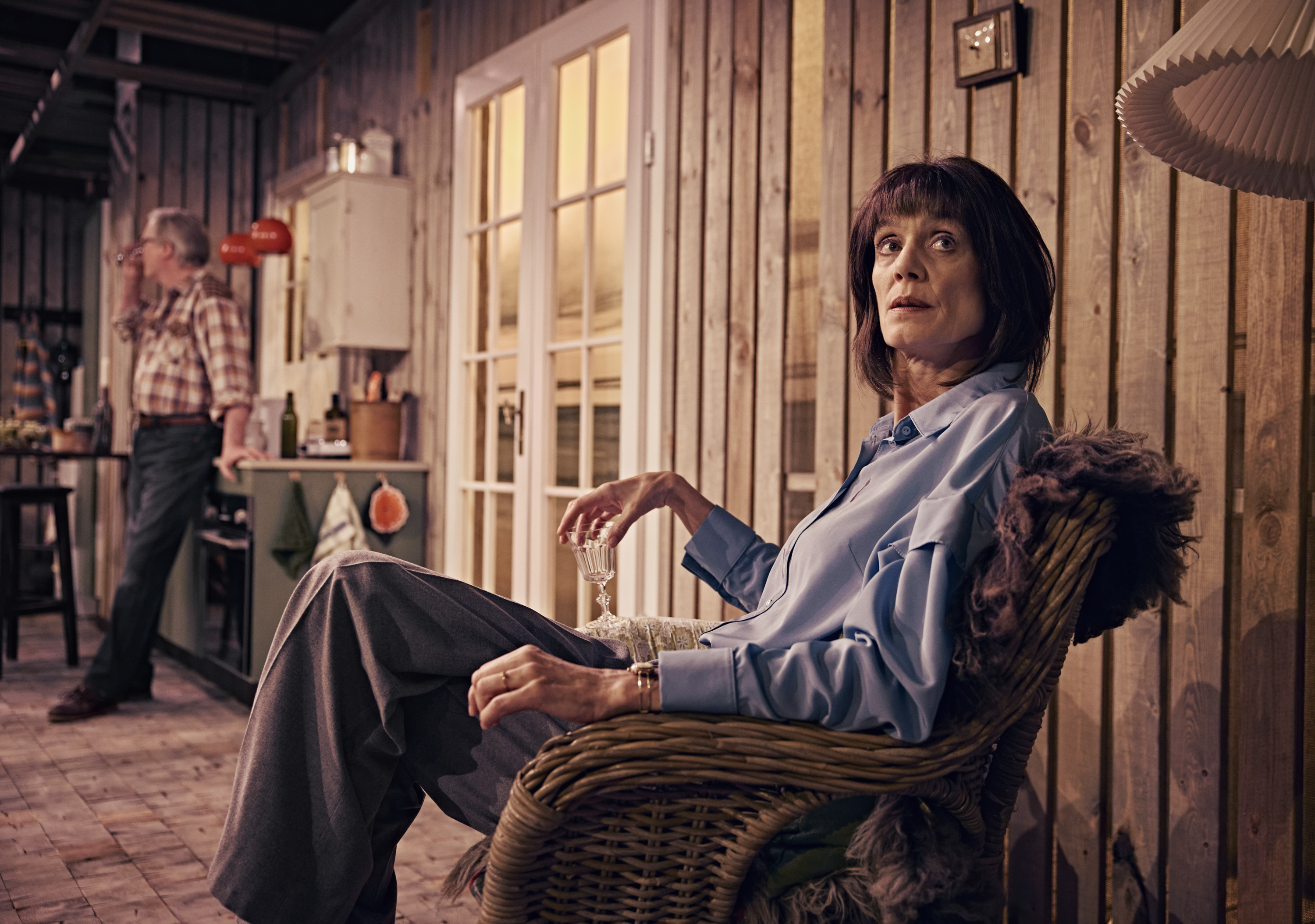 Kvinde sidder i stol. Holder et glas vin. Mand står længere væk lænet op ad køkkenbord. Fra teaterforestilling Børnene på Odense Teater.