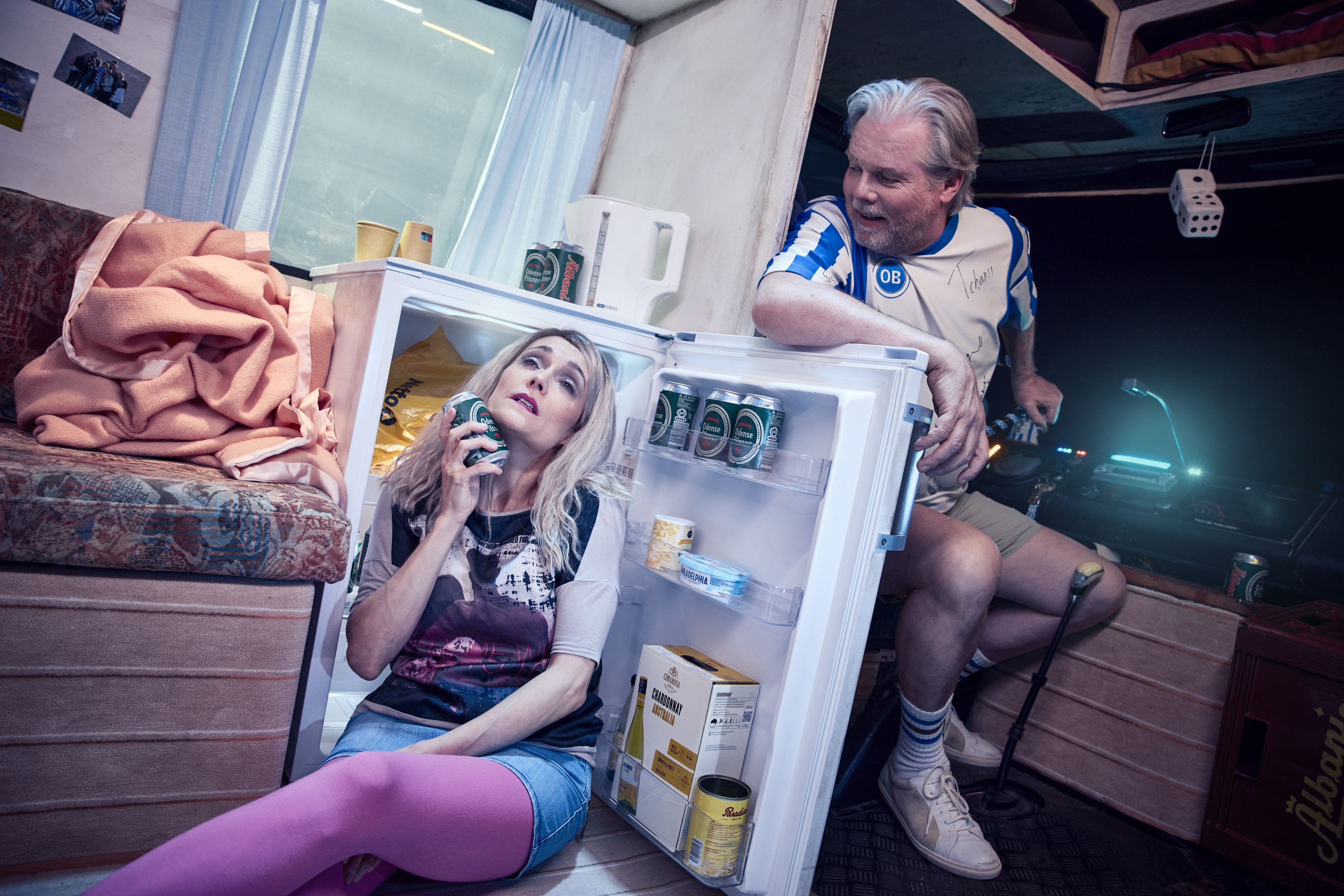 Kvinde sidder på gulvet foran lille kølskab og holder en dåseøl op mod ansigtet. En mand sidder og kigger smilende ned på hende. Fra teaterforestillingen Miraklet i Madrid på Odense Teater