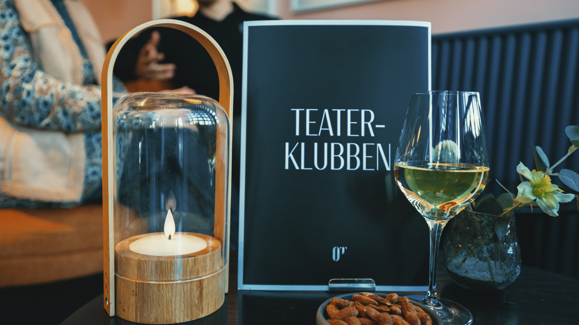 To mennesker i baggrund der sidder i en sofa. Et bord med stearinlys, et glas hvidvin, mandler i en skål, en blomst i vase, samt et skilt med skriften Teaterklubben og logo for Odense Teater.
