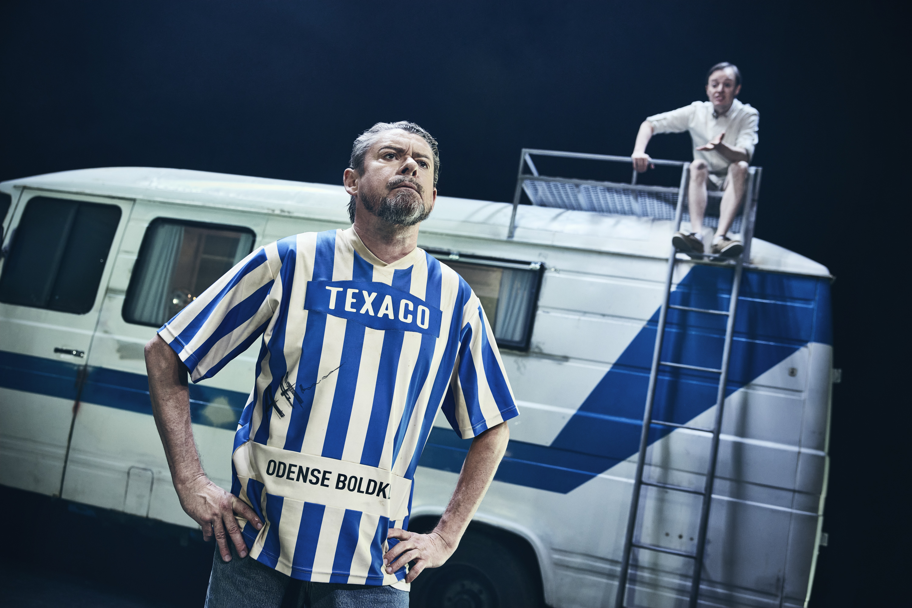 En mand i OB fodboldtrøje står foran en gammel autocamper. En ung mand sidder på taget. Fra teaterforestillingen Miraklet i Madrid på Odense Teater