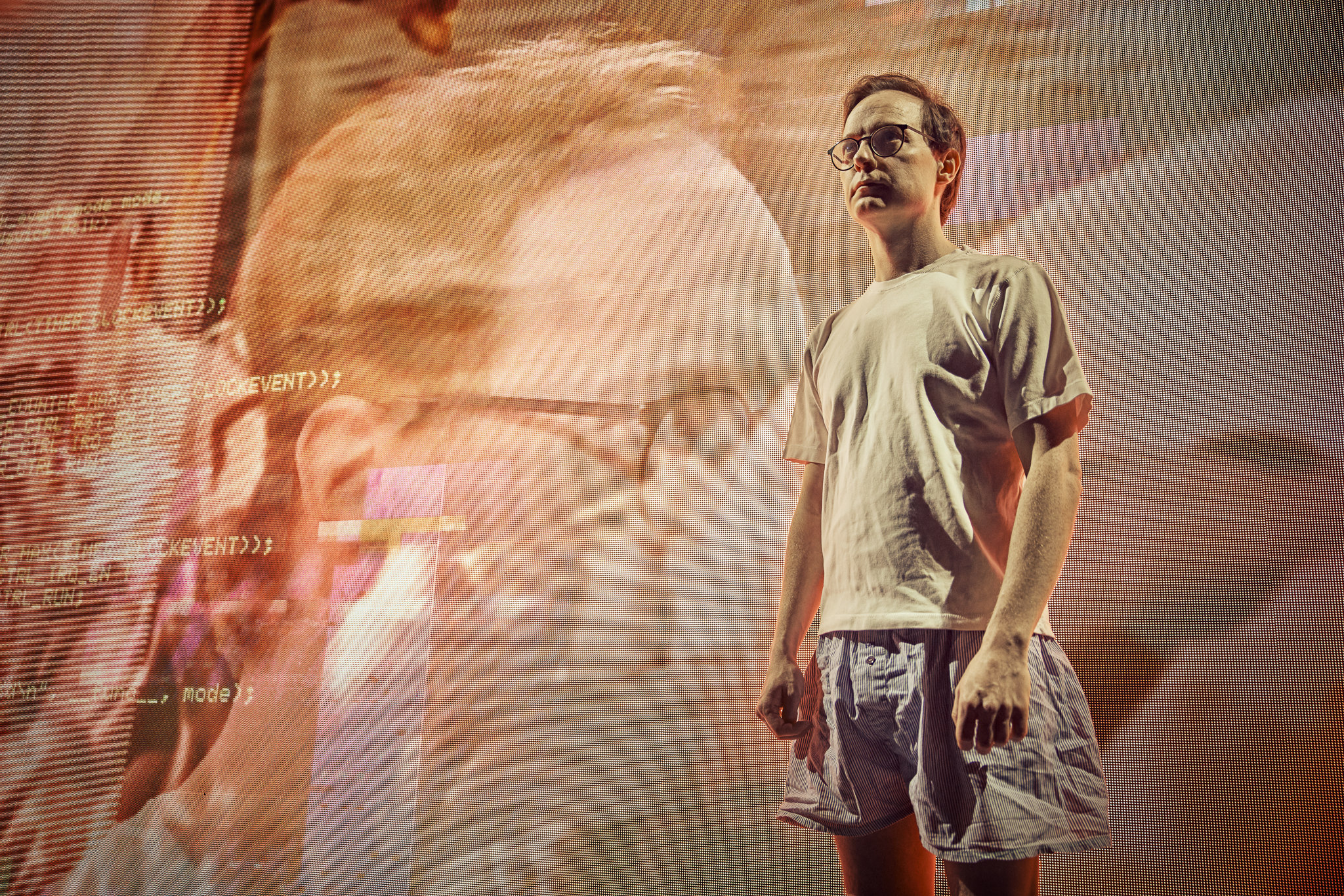 En mand i t-shirt og shorts står i et gyldent lys fra en storskærm bag ham. Fra forestillingen AI know you på Odense Teater