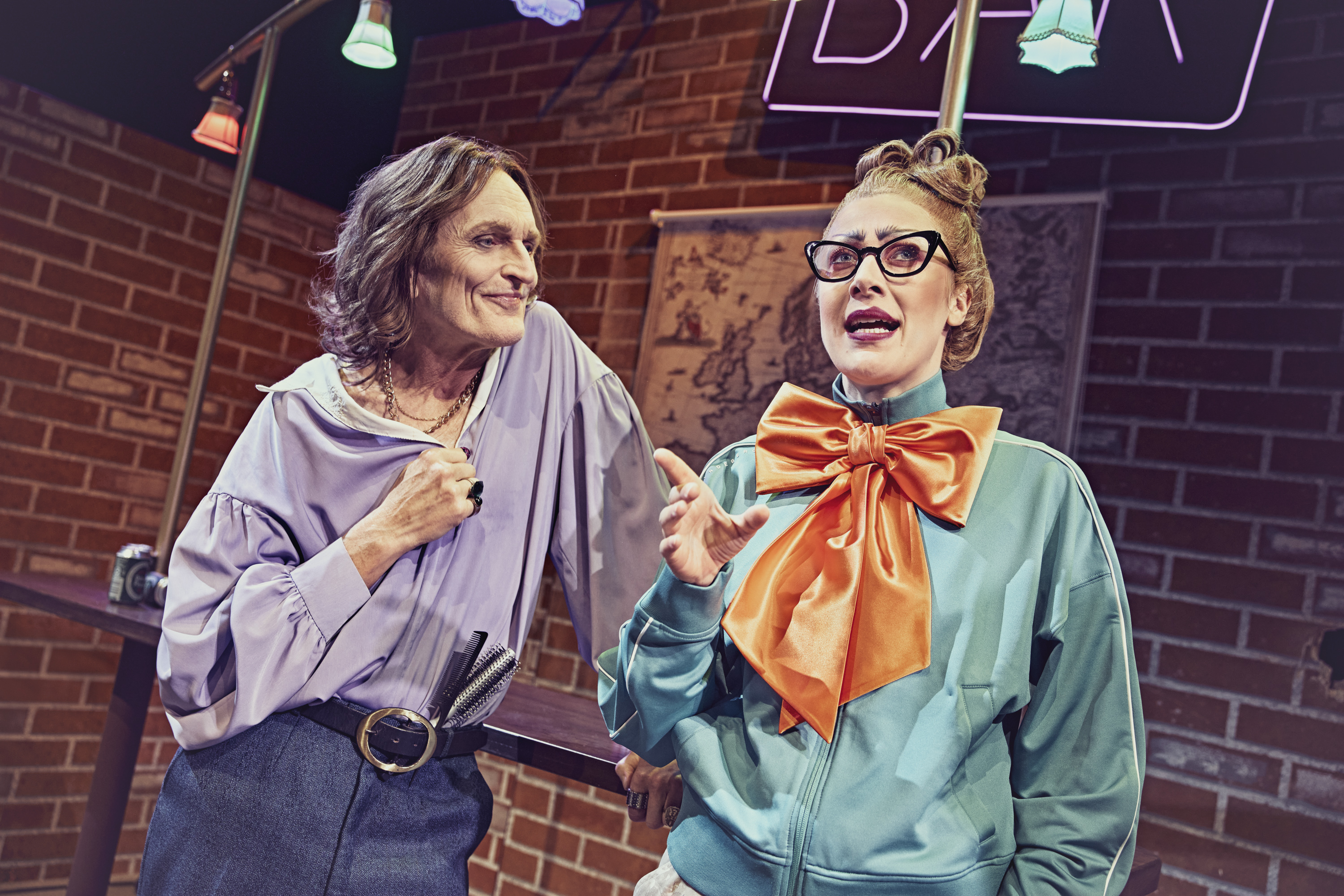 To personer står og taler sammen. Kvinden har briller og en meget stor orange sløjfe. Fra teaterforestillingen Den politiske kandestøber på Odense Teater