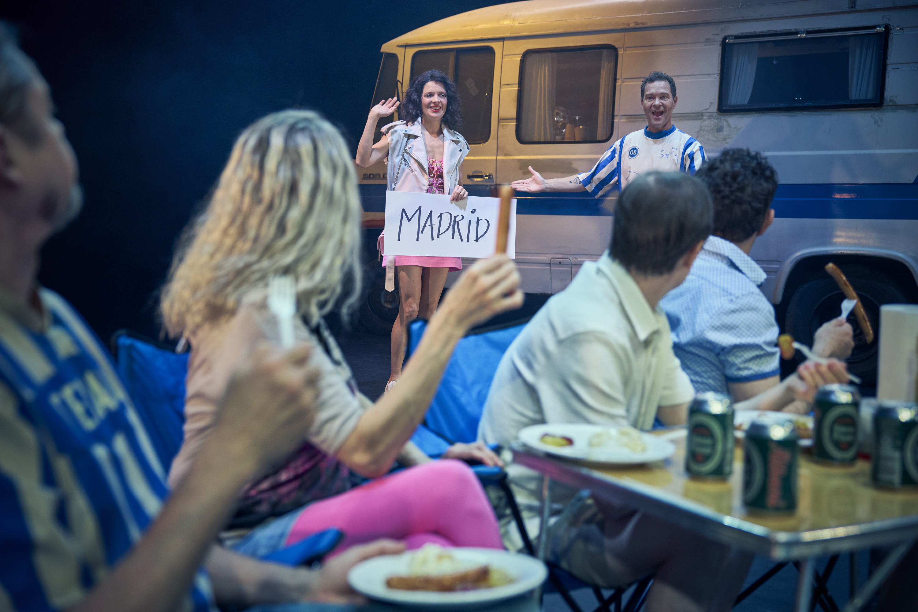 En kvinde står foran en autocamper med papskilt med teksten Madrid. Flere mennesker sidder ved campingbord og spiser. Fra teaterforestillingen Miraklet i Madrid på Odense Teater