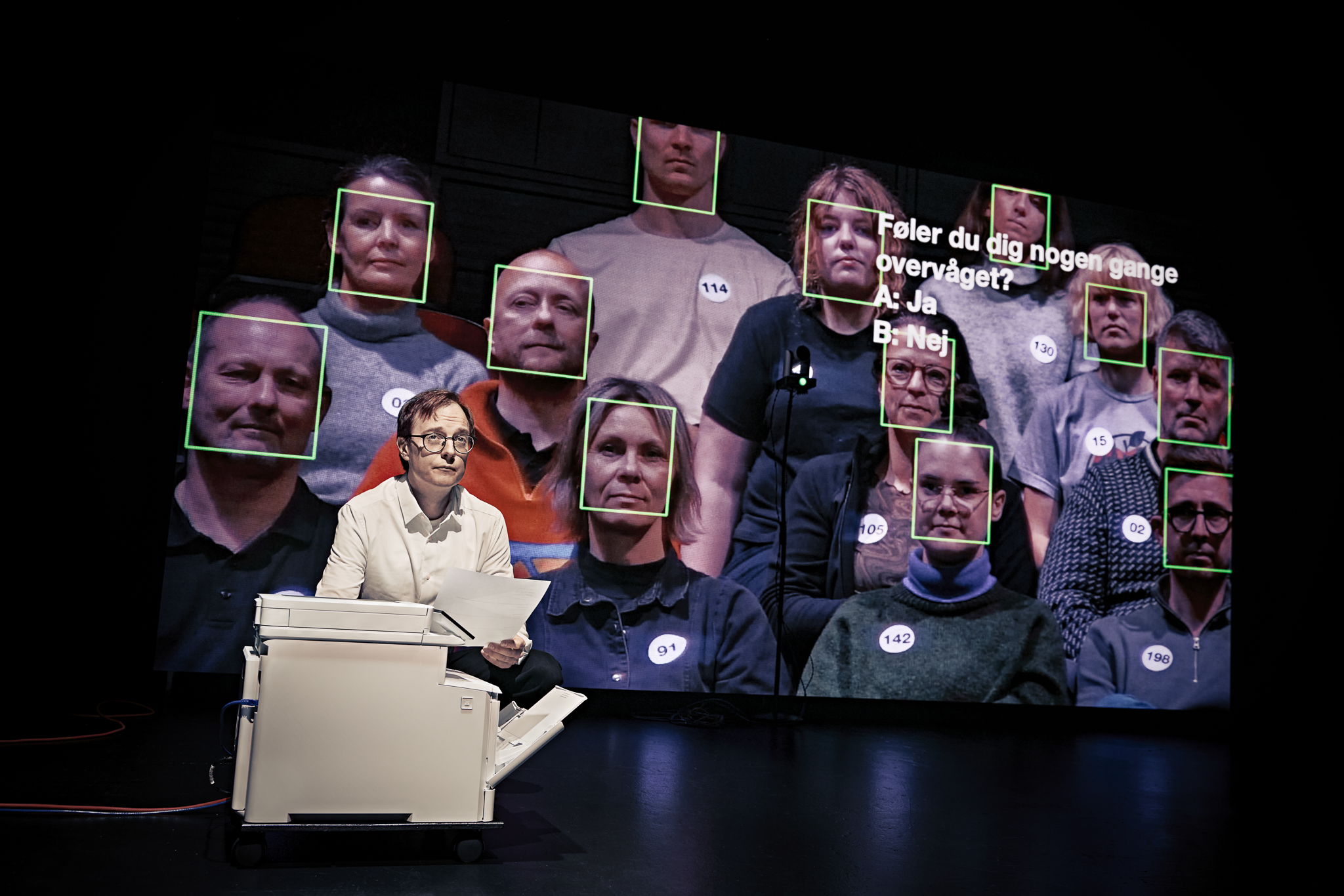 En mand i hvid skjorte sidder på hug foran en printer. Bag ham er en stor skærm med 12 mennesker hvor der der er en grøn firkant rundt om hvert ansigt. Fra forestillingen AI know you på Odense Teater