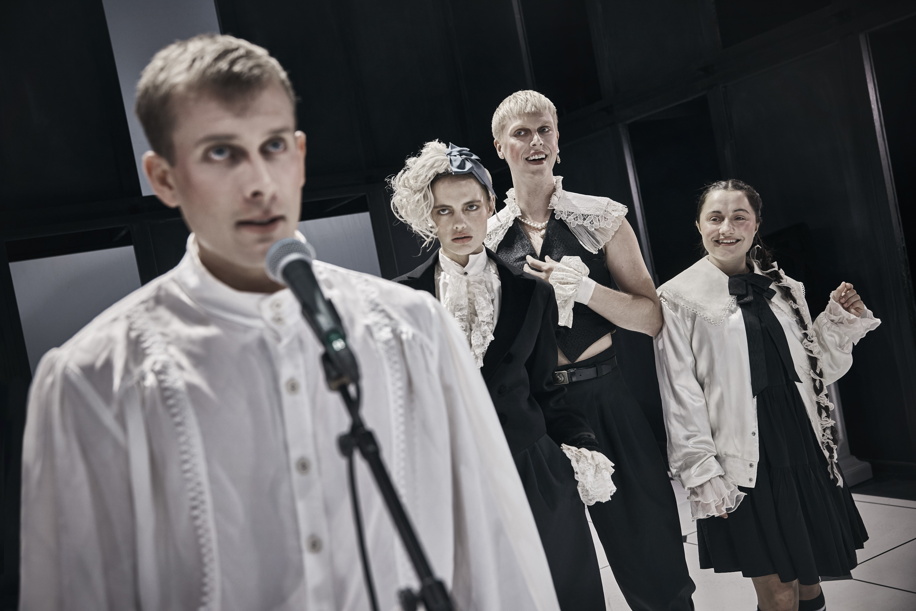 Ung mand i hvid skjorte står på scenen foran mikrofon. Tre tilhører stå i baggrunden. Fra forestilling Cyrano de Bergerac på Odense Teater