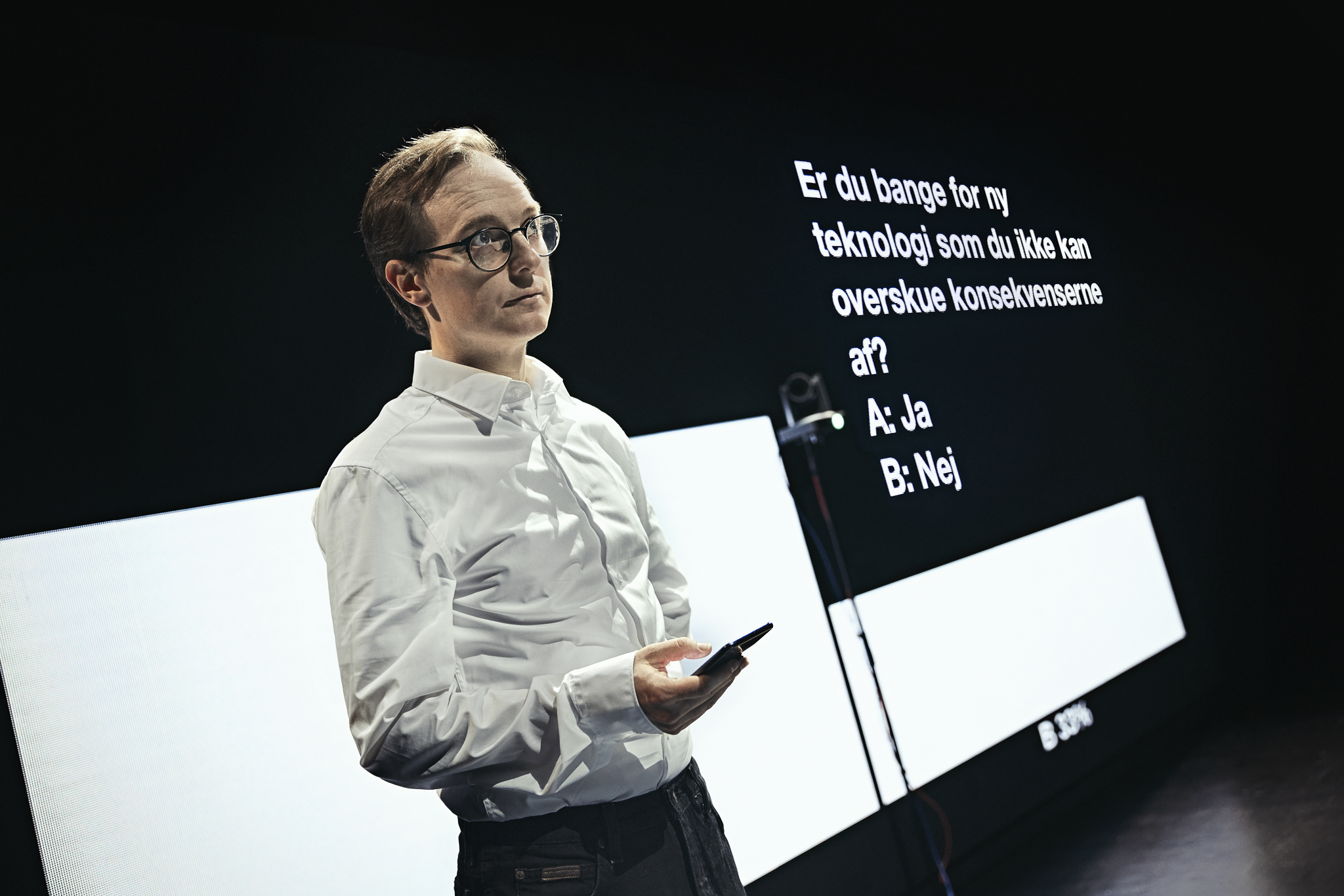 En mand i en hvid skjorte står med en tlf. i hånden foran en skærm med diagram og tekst. Fra forestillingen AI know you på Odense Teater
