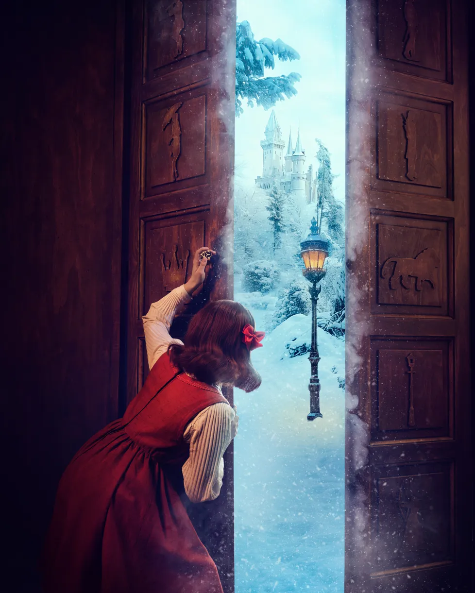 Narnia på Odense Teater. Lille pige i rød kjole og hvid bluse med rød sløjfe i håret kigger ind gennem døråbning på et magisk skab. Landskabet derinde er snefyldt og et slot og gammel lygtepæl.
