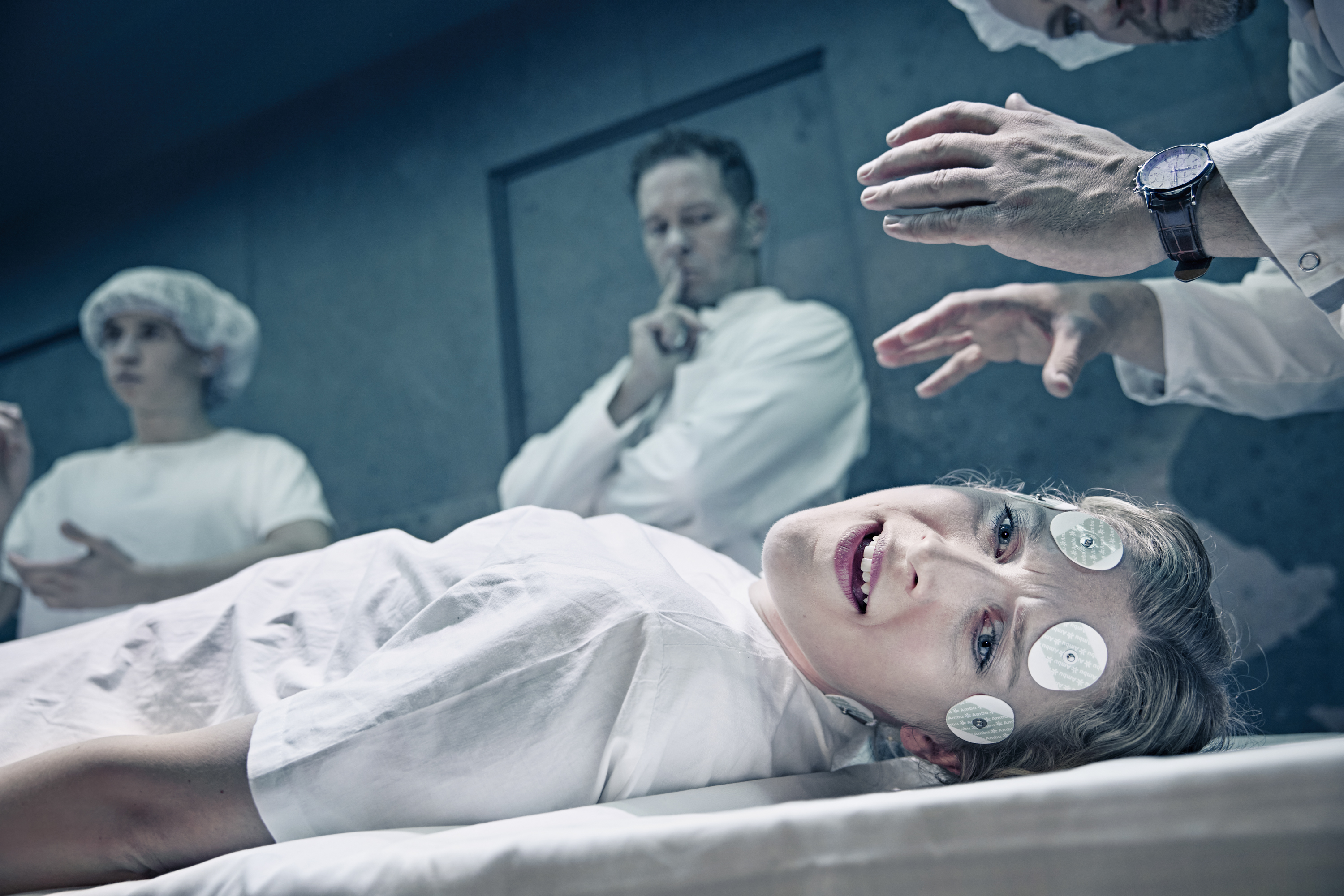 En kvinde ligger på et leje. Hun har elektroder i panden. Læger står i baggrunden i musicalen Next to Normal på Odense Teater