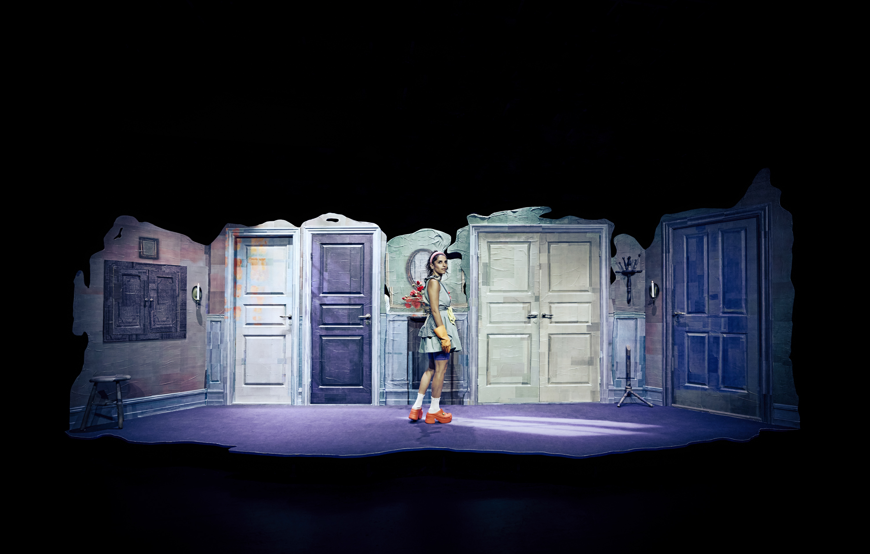 Kvinde i kort kjole og orange sko står på scene i en scenografi med med mange døre. Mørkt billede med enkelte farvepletter på døre og tøj. 
