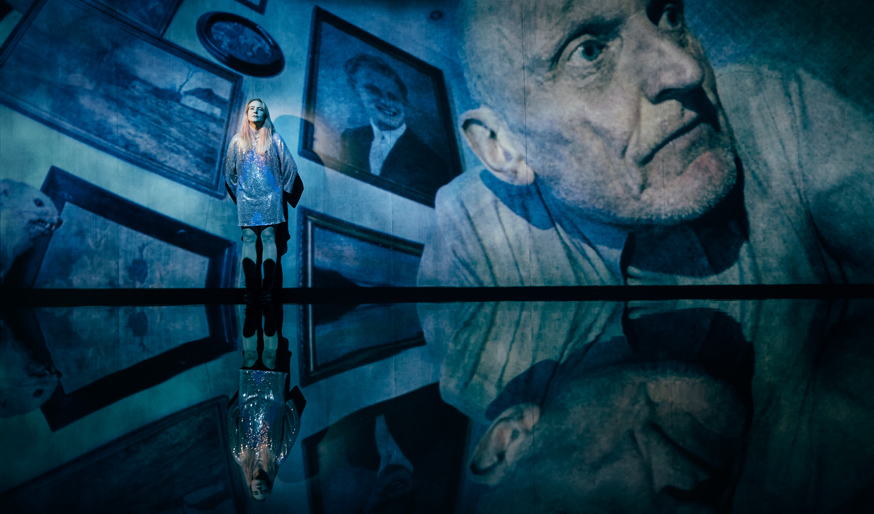 Ung kvinde i pailetkjole står op ad stor skærm, hvor der et ansigt af ældre mand. Fra forestilling Maria på Odense Teater.