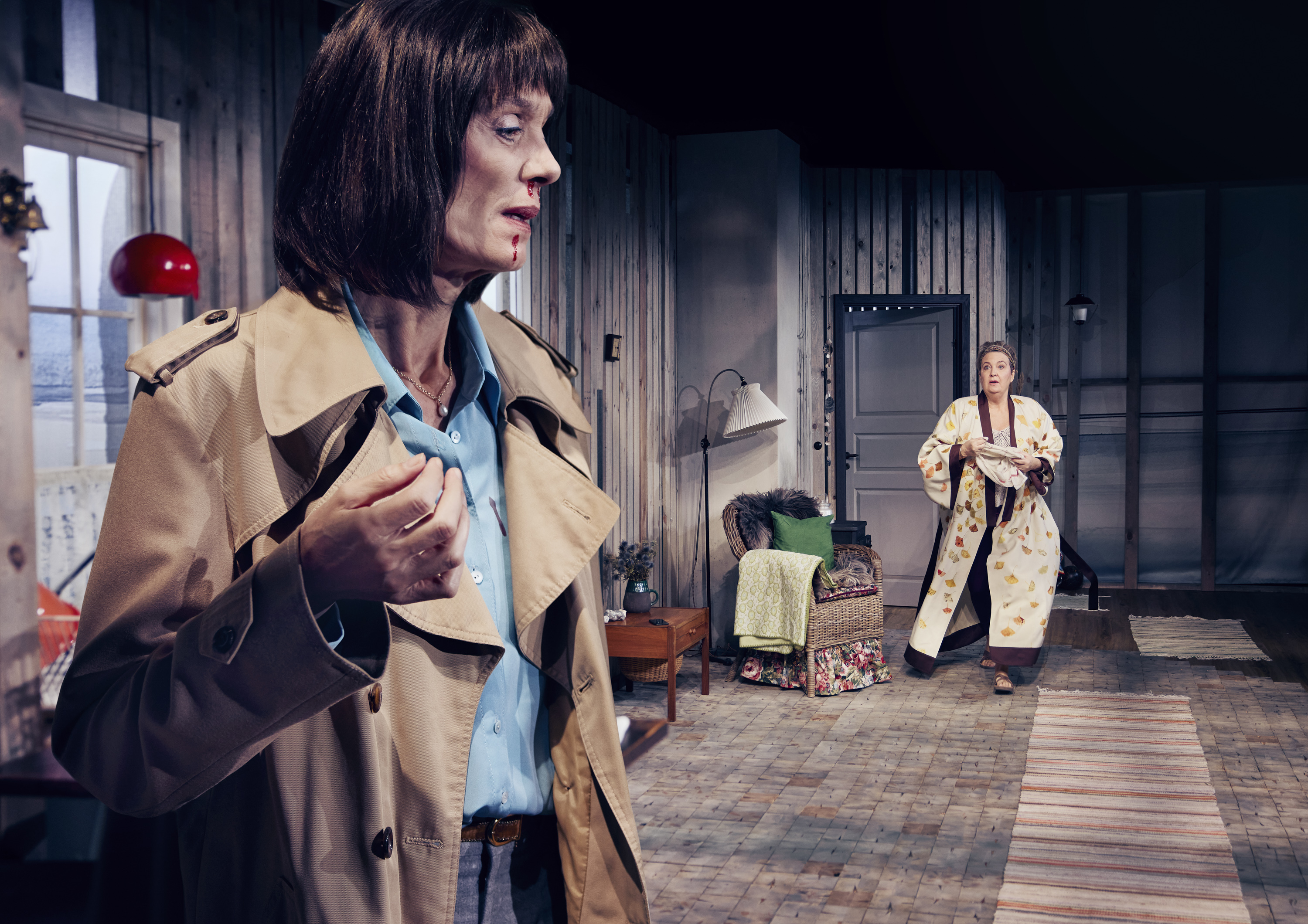 Kvinde i trenchcoat står i forgrund. Hun har næseblod. Kvinde i kimono står i baggrunden. Fra teaterforestilling Børnene på Odense Teater.
