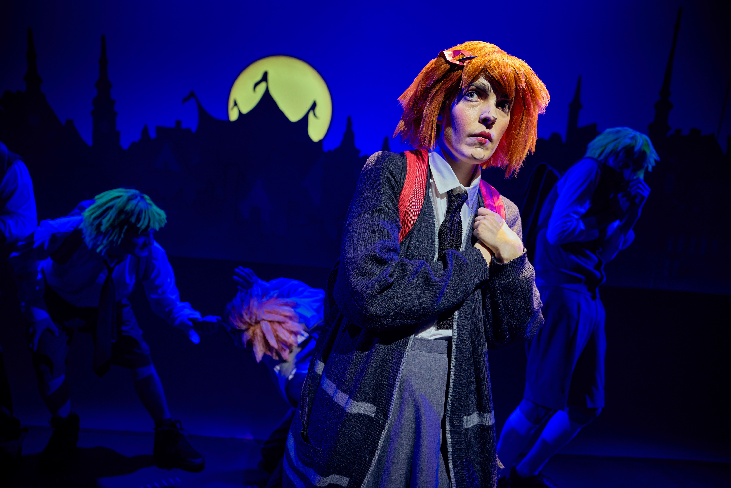 En pige med orange hår og skoleuniform skutter sig. Personer i baggrund som driller hende. Fra forestillingen Håndbog for superhelte på Odense Teater