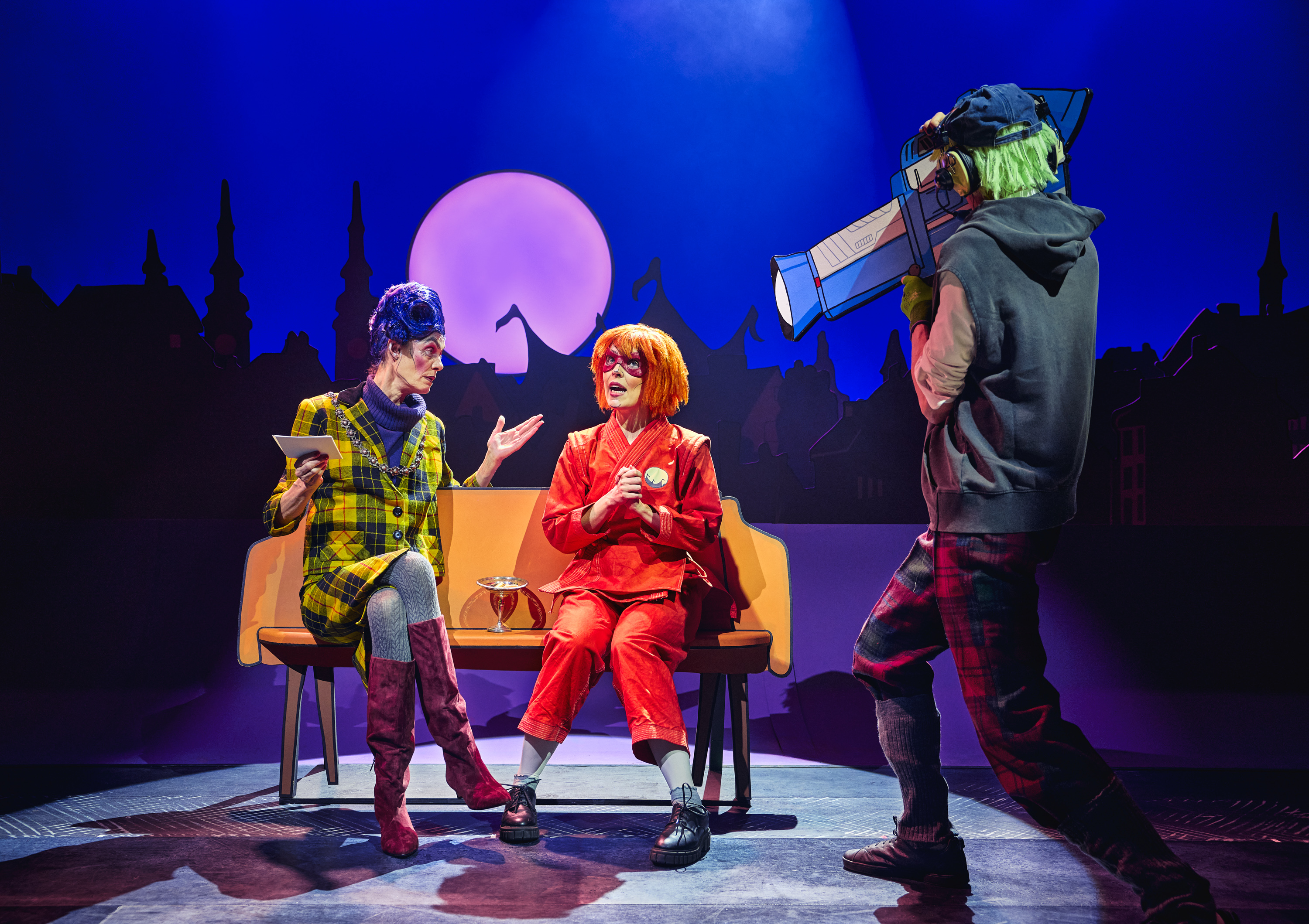 Pige med rødt tøj, orange hår og rød heltemaske giver interview til kvinde med blåt hår. En kameramand i forgrunden. Fra forestillingen Håndbog for superhelte på Odense Teater