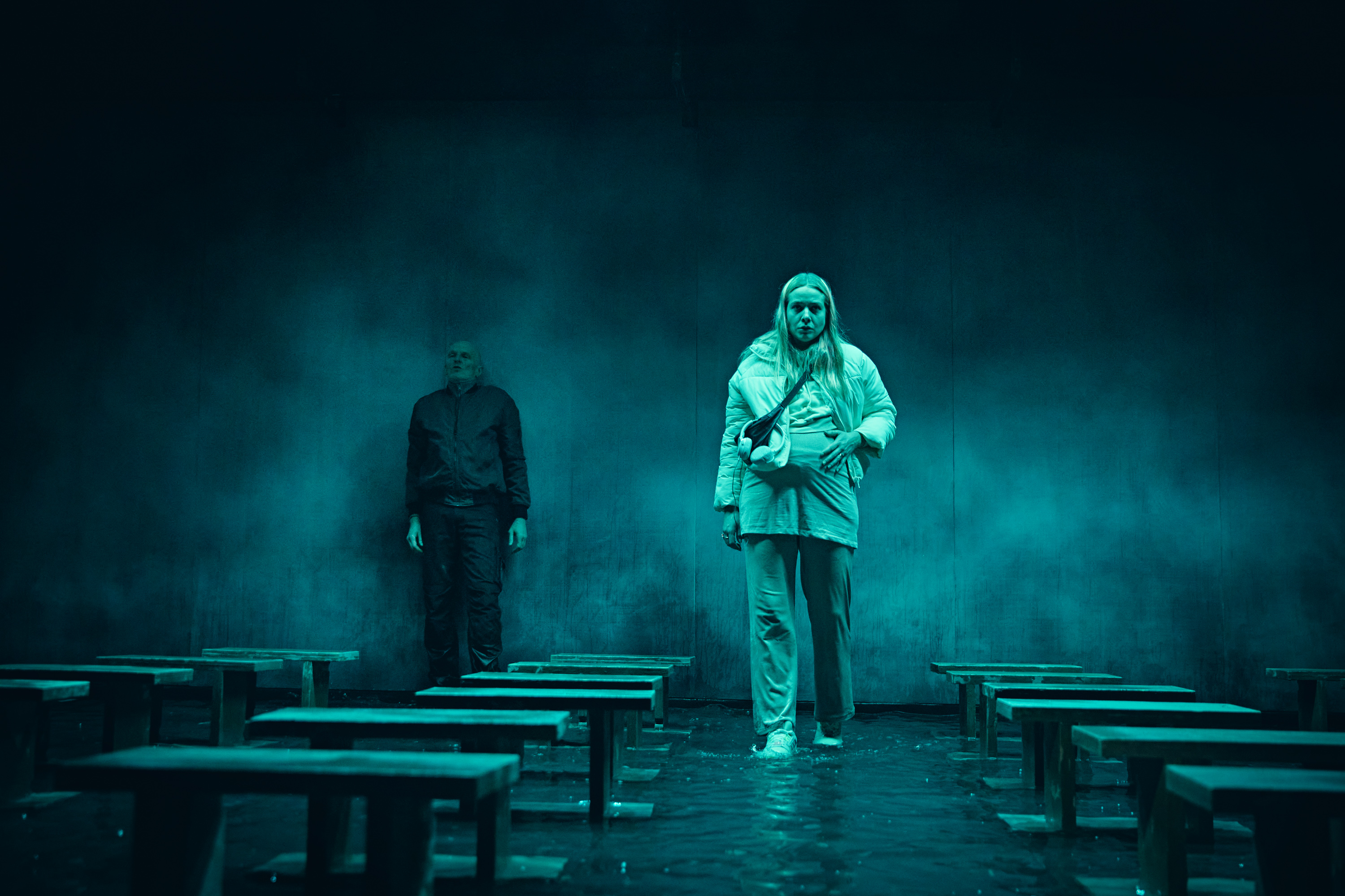 Ung gravid kvinde går på scene dækket af vand. Der står en mand i baggrund. Fra forestilling Maria på Odense Teater.