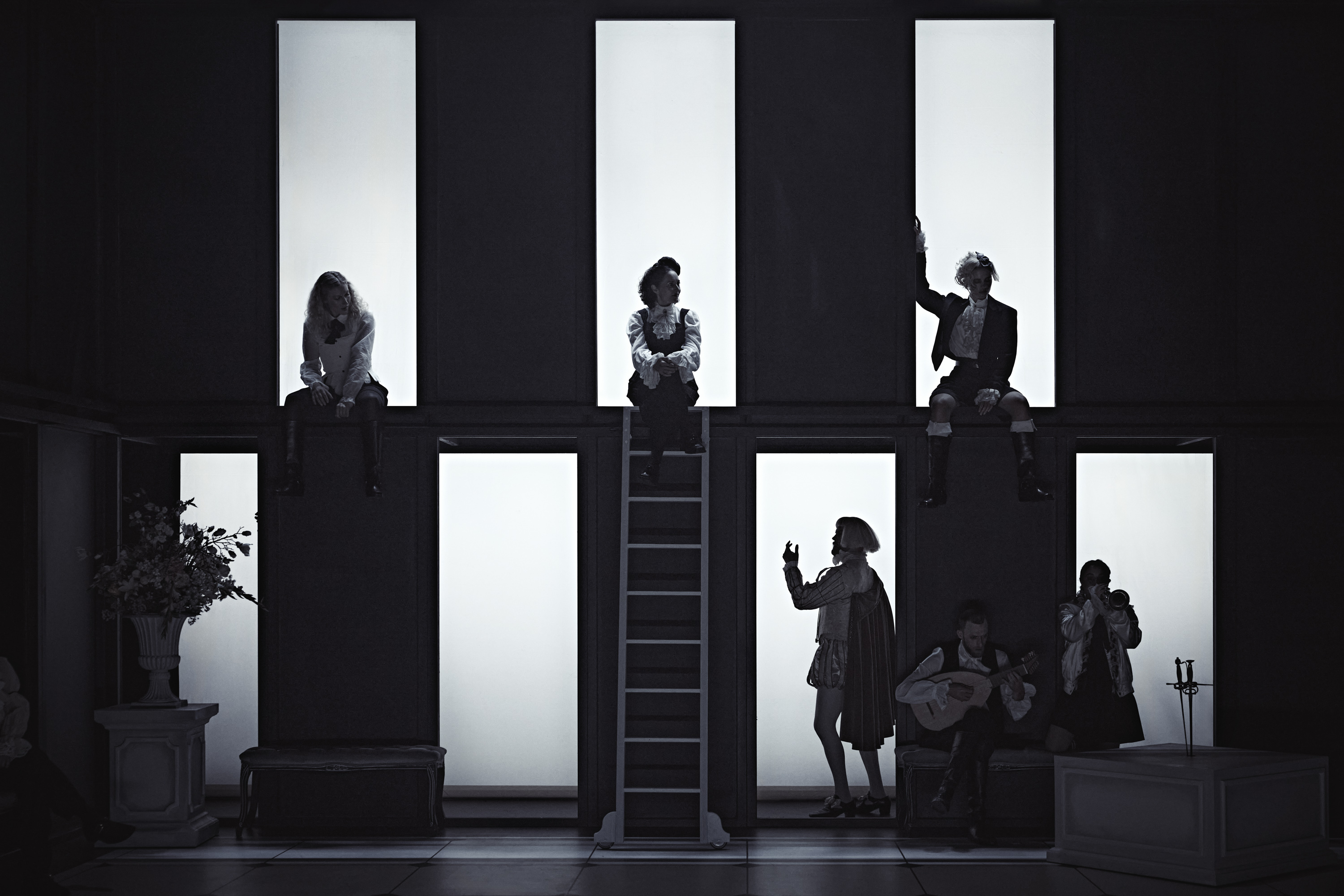 6 skuespillere på scene iført tøj i barokstil. En spiller på guitar. Tre sidder oppe i vinduer over de tre andre der er på gulvet. Sorte og hvide felter.