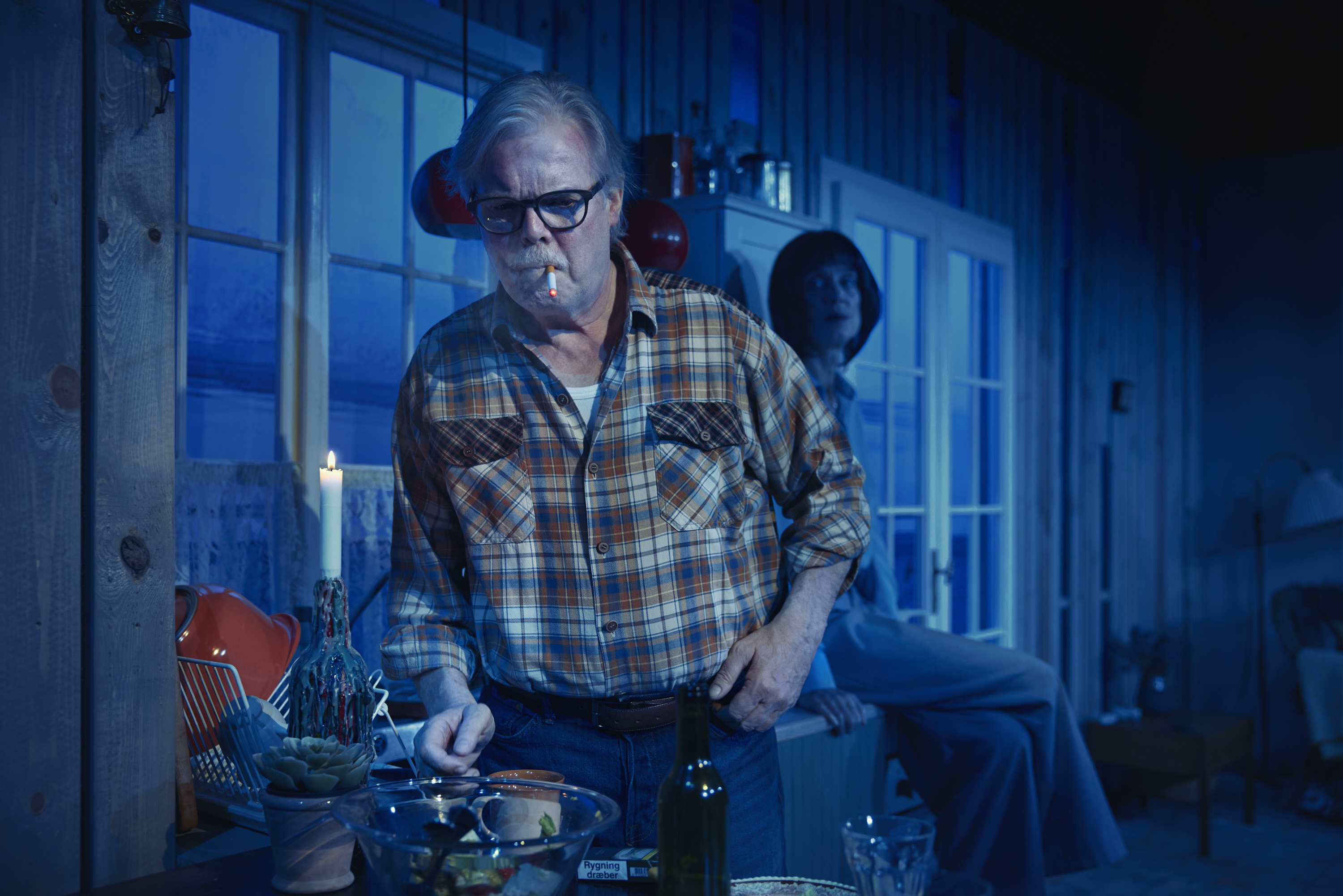 Mørkt billede. Mand i ternet skjorte ryger på cigaret. Kvinde sidder på et køkkenbord i baggrunden. Fra teaterforestilling Børnene på Odense Teater.