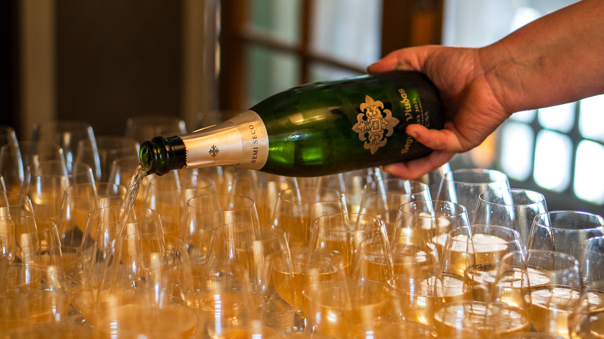 En hånd holder champagneflaske hen over mange glas. Fylder vin i glas. 